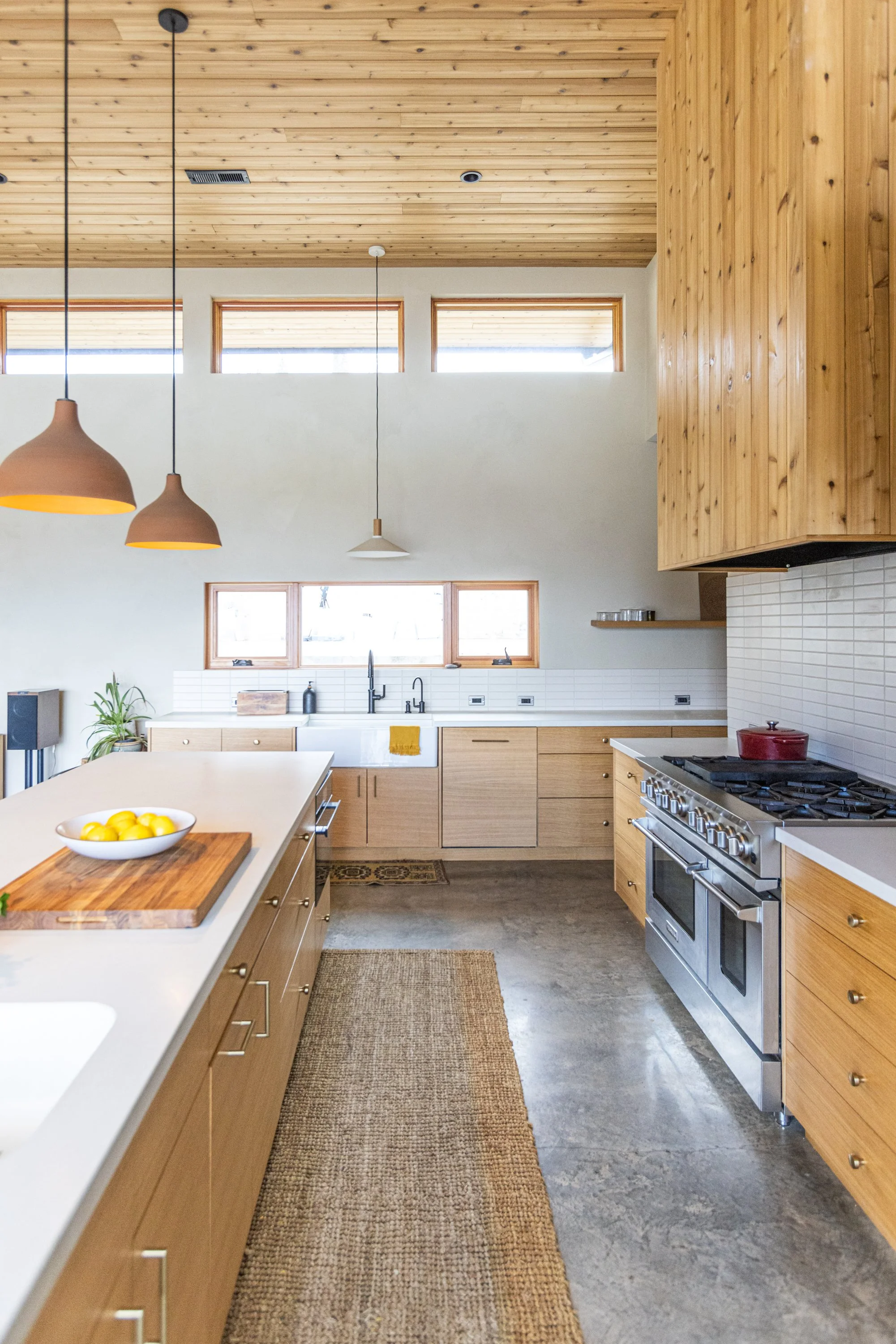 Modern kitchen with wooden cabinets, a kitchen island with a bowl of lemons, pendant lights, large windows, a stainless steel stove, and white tiled backsplash.