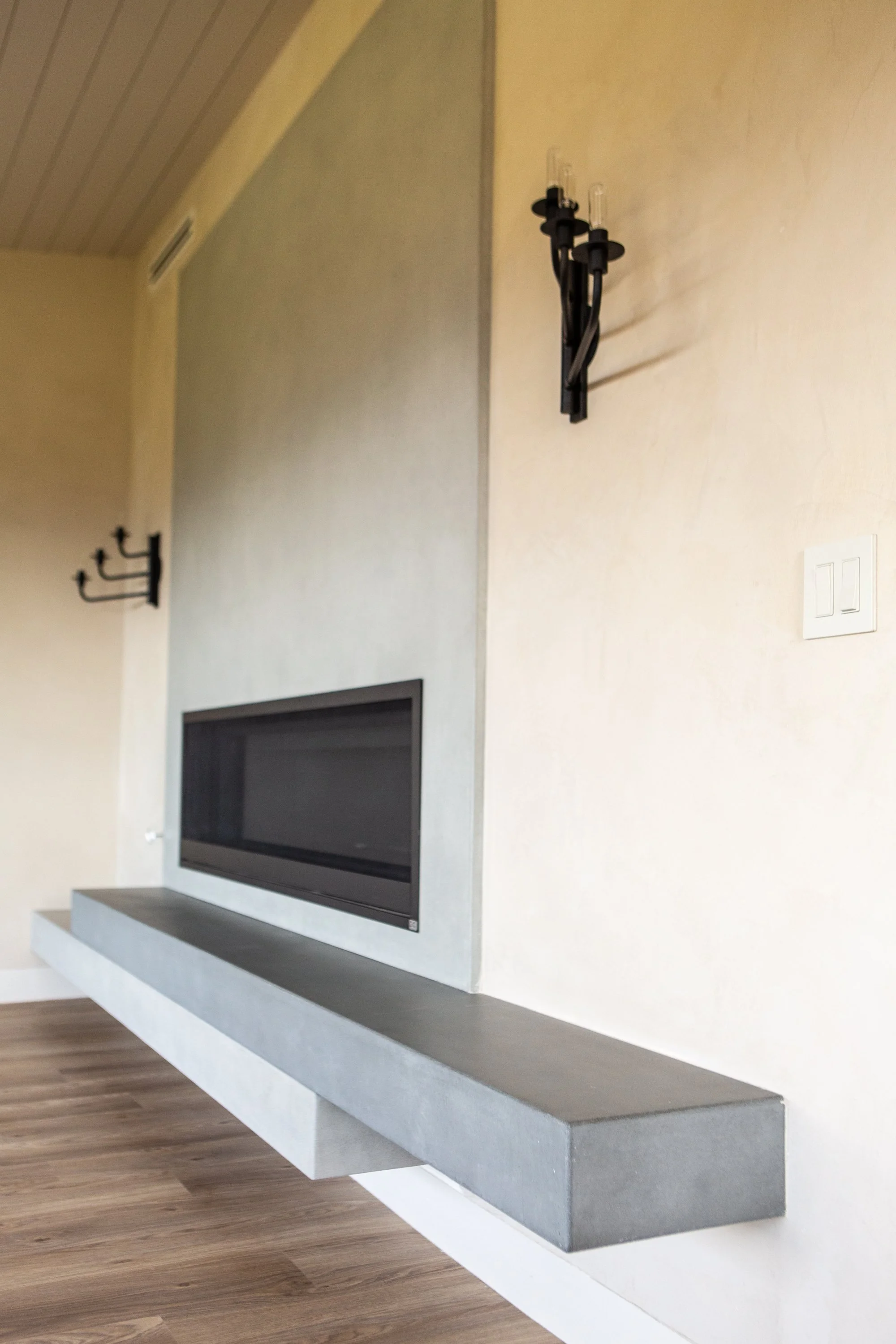 Modern living room corner with flat-screen TV mounted on the wall, white and gray built-in fireplace, and minimalistic decor. Wall sconce with candles on the right wall, wood floor, and a floating gray shelf.