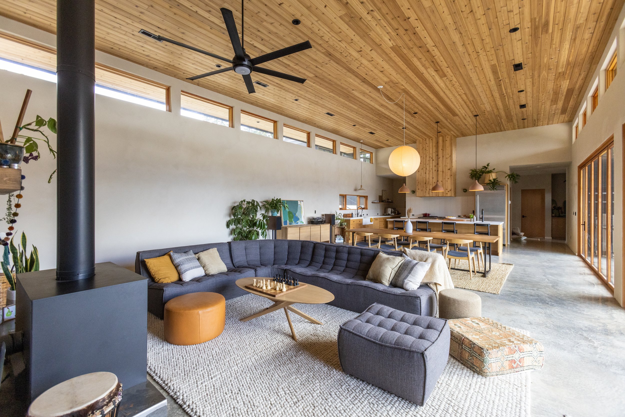 Open living room with wood-paneled ceiling, black ceiling fan, large windows, gray sectional sofa with pillows, indoor plants, dining table with chairs, lamp, and kitchen in the background.