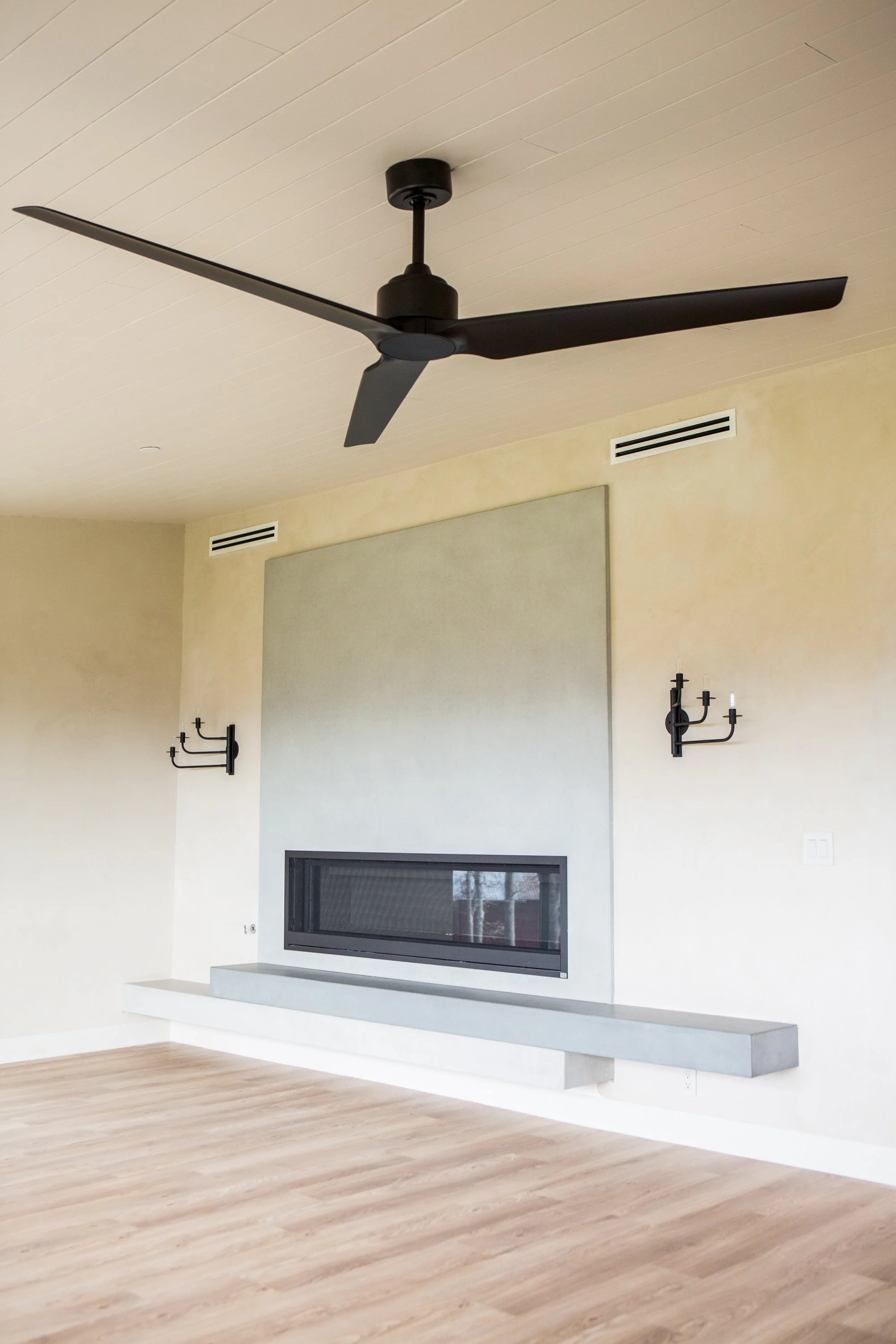 Living room with light-colored walls, a modern black ceiling fan, a wall-mounted fireplace, and a large window, wooden flooring, and minimalistic wall sconces.