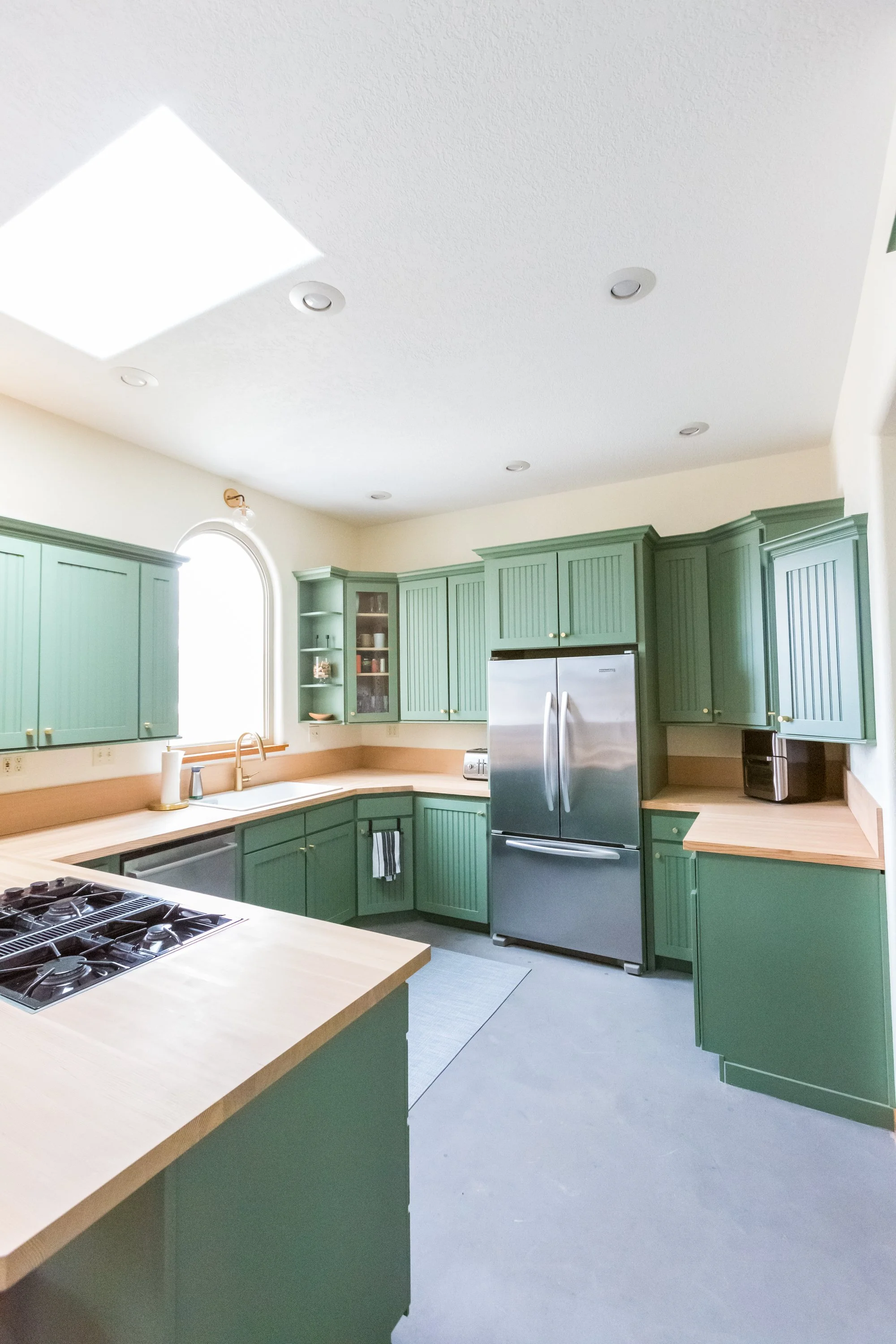 Kitchen with green cabinets, stainless steel refrigerator, beige countertops, and a gas stove with four burners. A window with an arched top is above the sink area, with sunlight coming through.