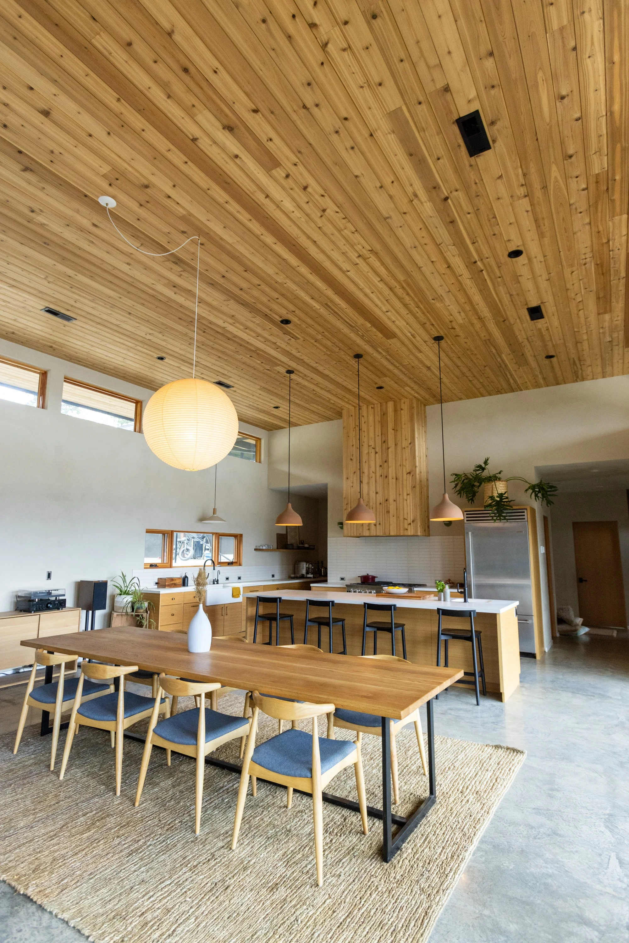 Open-concept kitchen and dining area with wooden ceiling and furniture, pendant lighting, and a textured rug under a dining table with chairs.