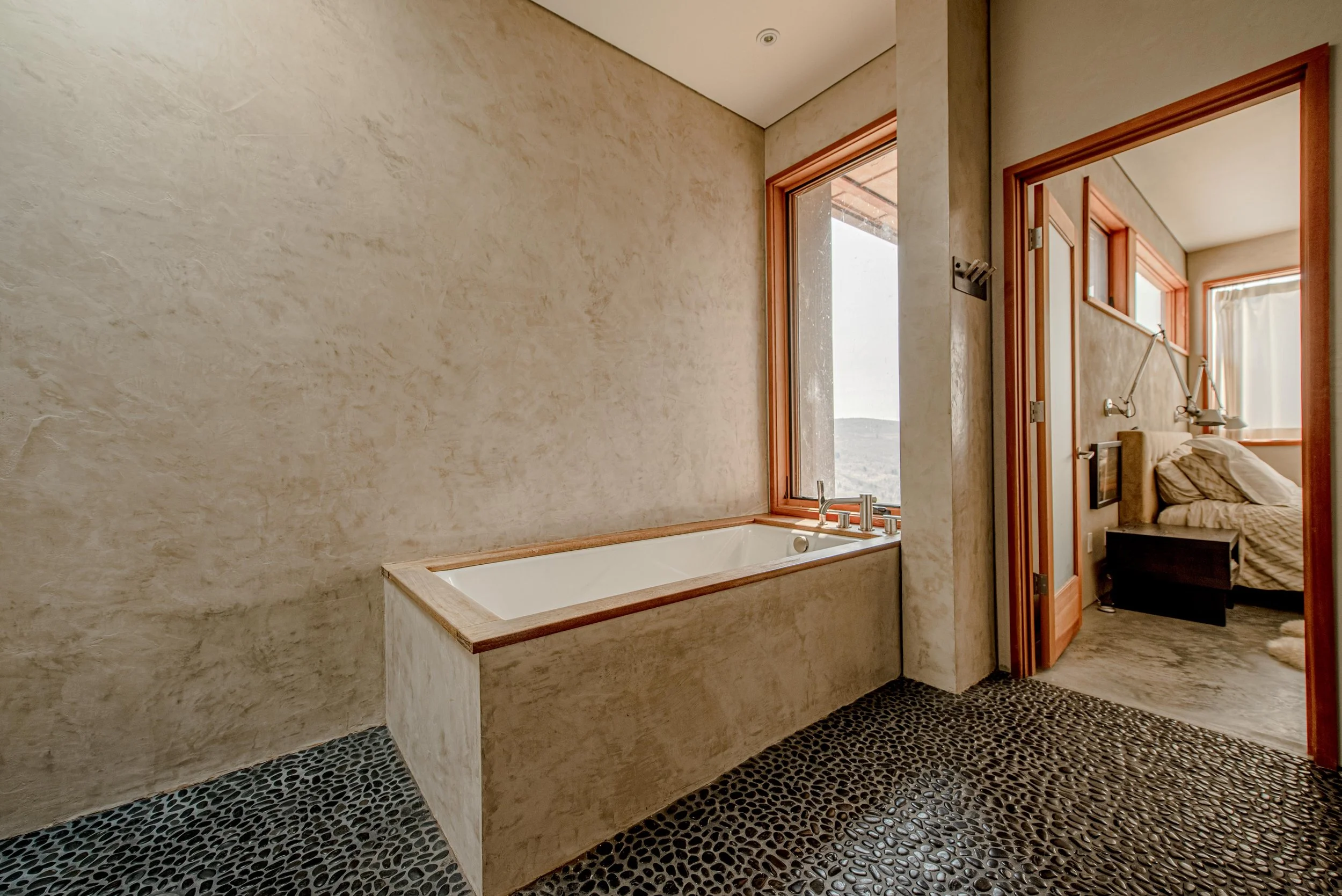 Bathroom with a bathtub, large windows, and a bedroom with a bed and window visible through open doors.