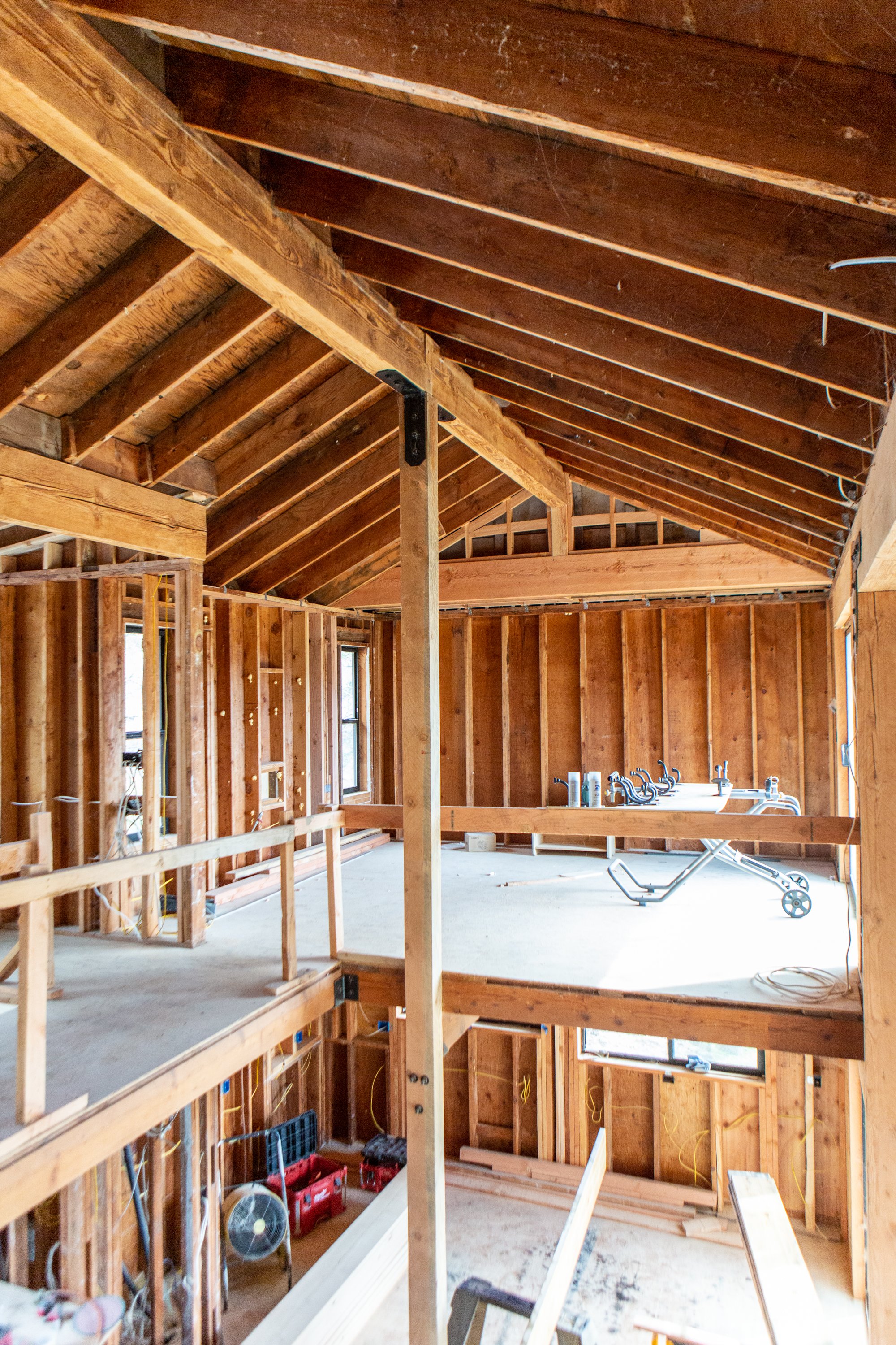 Interior view of a house under construction, showing exposed wooden framing and roof trusses, with some construction tools and supplies.