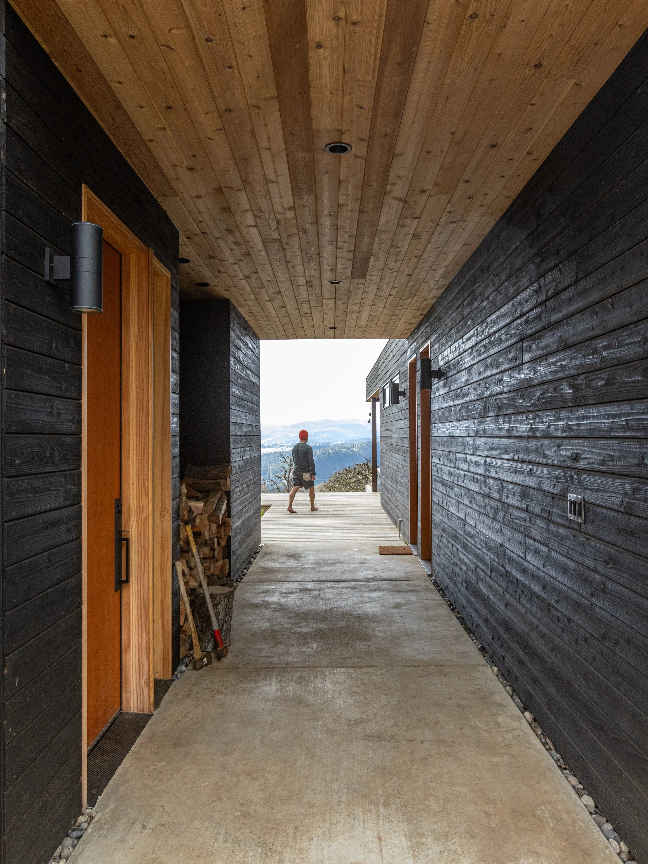 View through an outdoor hallway with black wooden walls, a natural wood ceiling, and a concrete floor. A person wearing a red cap and shorts is standing outside in the distance overlooking a landscape with trees and hills.