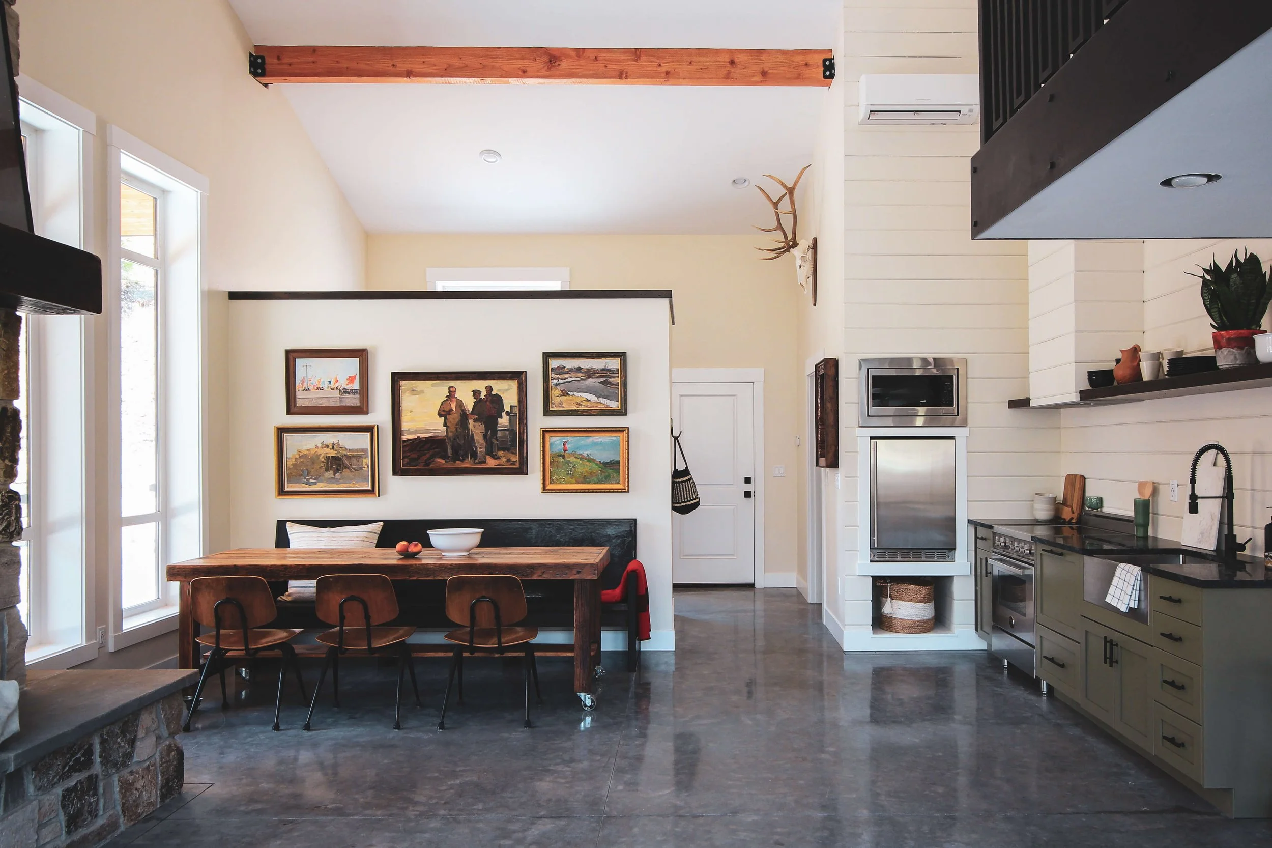 Open-concept kitchen and dining area with white panel walls, a wooden dining table, six chairs, a gallery wall with framed artwork, and kitchen appliances including a stainless steel oven and microwave, with decorative plants and an antler wall mount