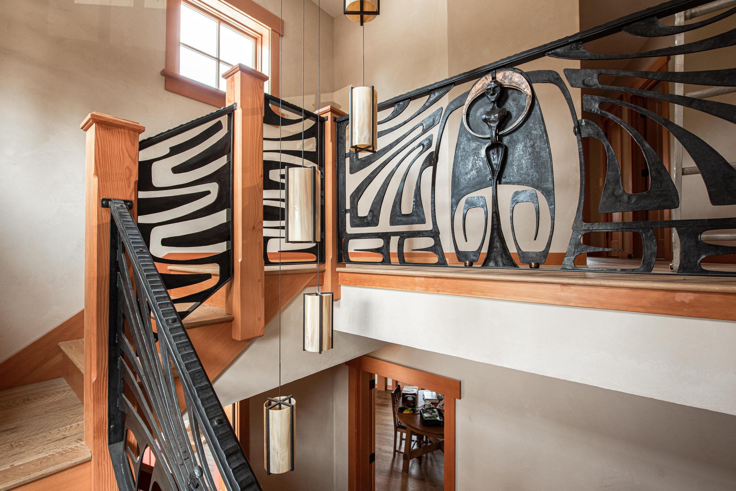 Interior view of a staircase with black metal railings featuring abstract and organic shapes, wooden posts, hanging cylindrical lights, a high beige wall, and a window letting in natural light.