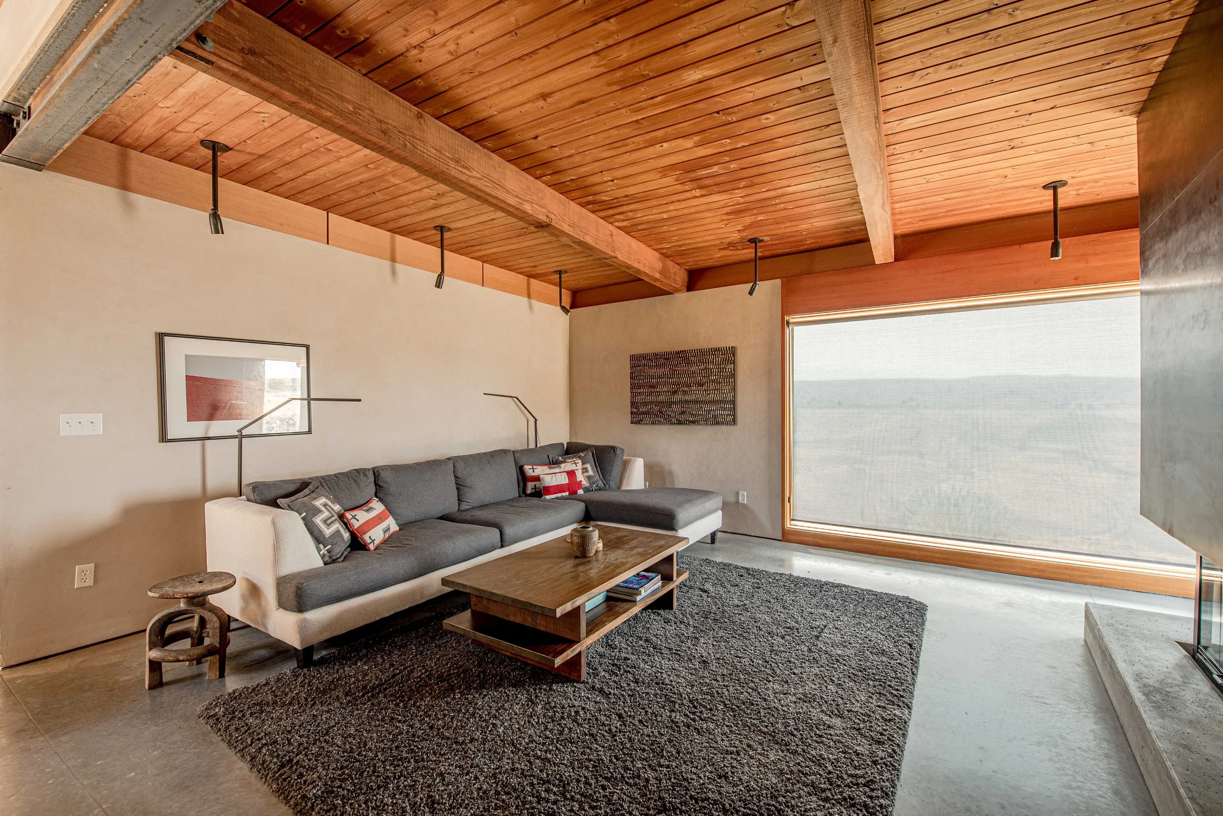 Living room with a gray sofa, wooden coffee table, and a black area rug. The ceiling is made of wood planks with exposed beams and ceiling lights. Large window with a beige roller shade.