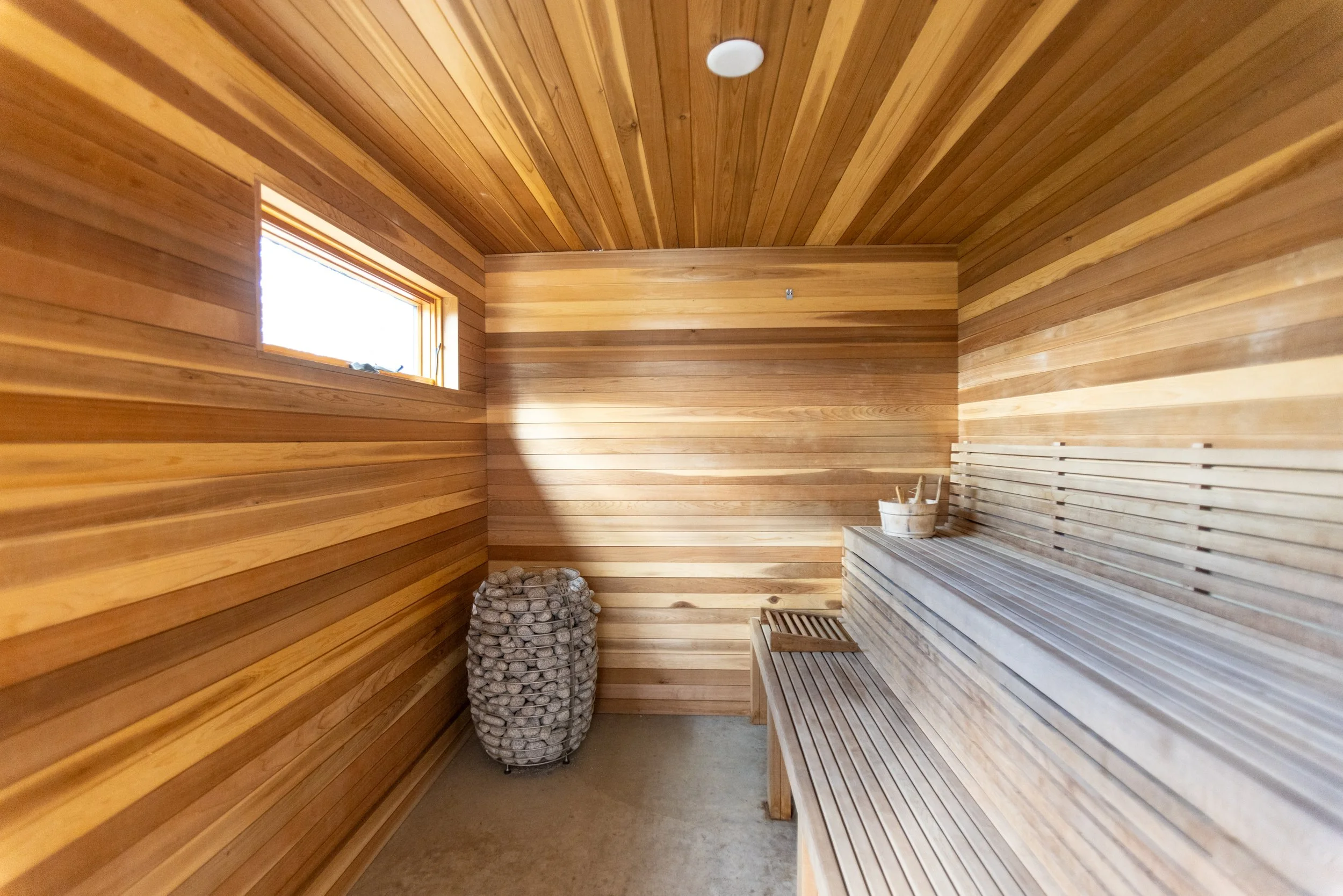 A sauna room with wooden walls and ceiling, a window, a bench, a bucket of stones, and a sauna ladle.
