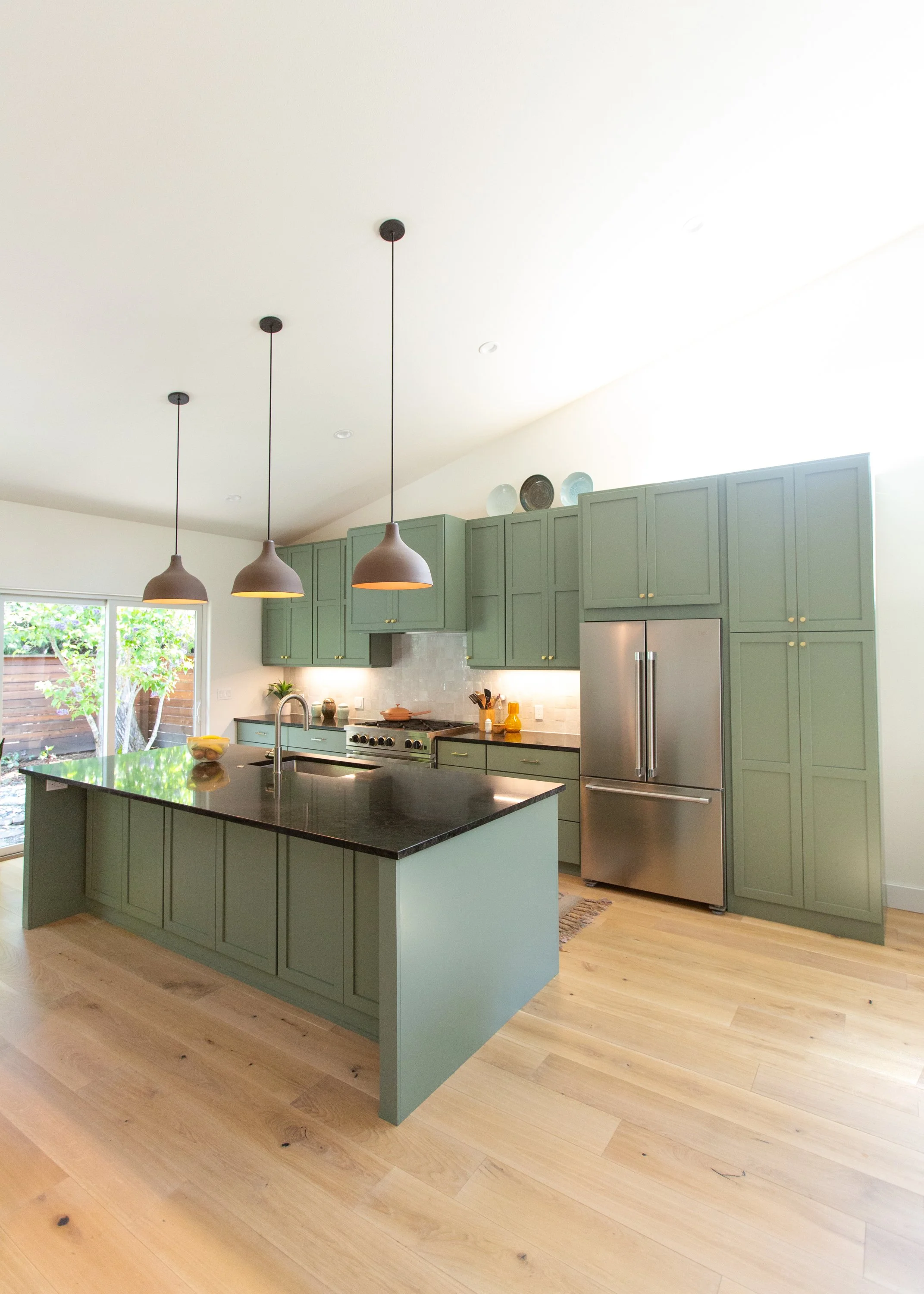 Modern kitchen with green cabinetry, black island countertop, stainless steel refrigerator, and hanging pendant lights.