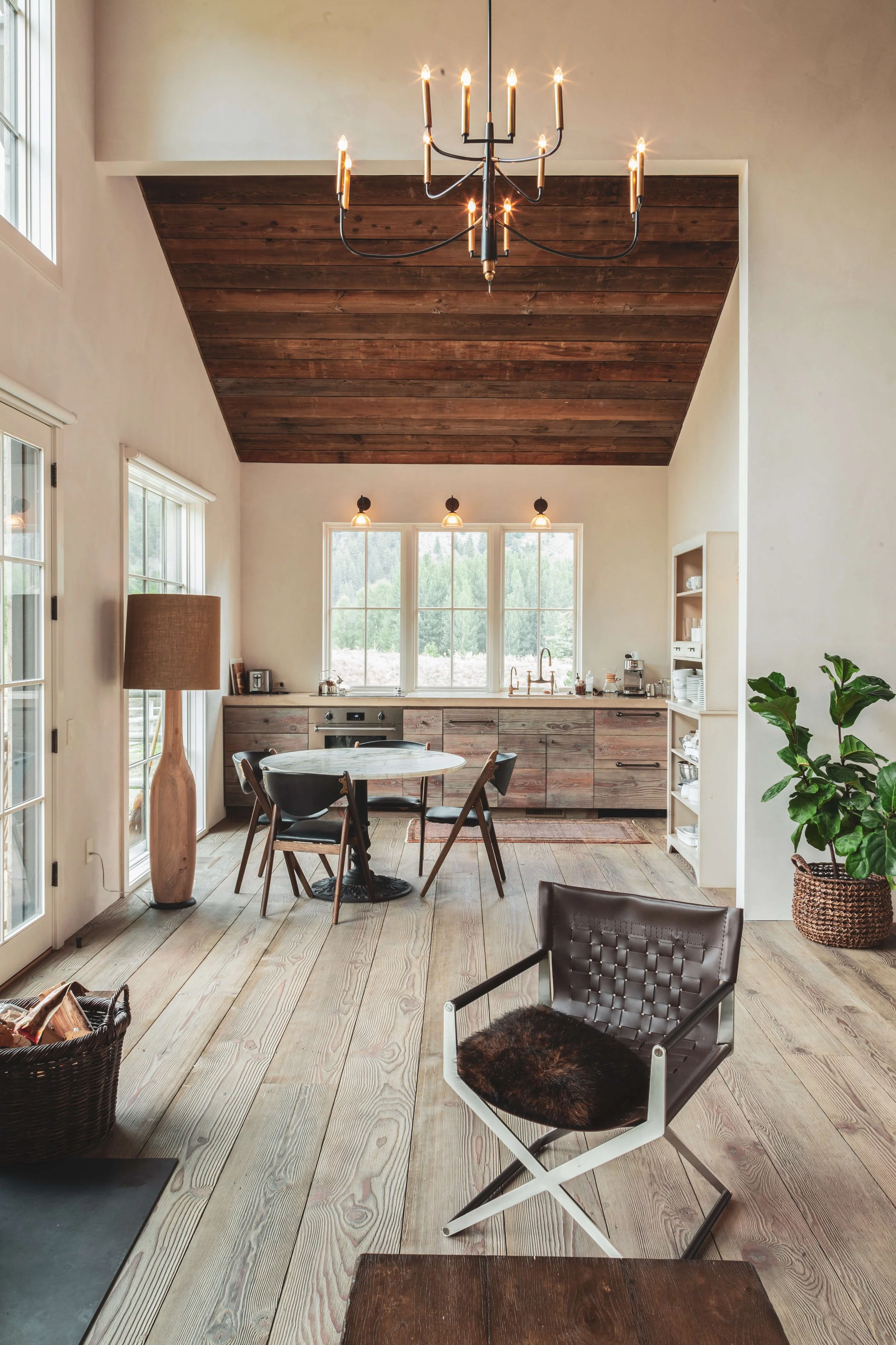 A cozy kitchen and dining area with a wooden high ceiling, a black chandelier, and natural light from large windows. The space features wooden floors, a round dining table with black chairs, a kitchen counter with cabinets, and houseplants.