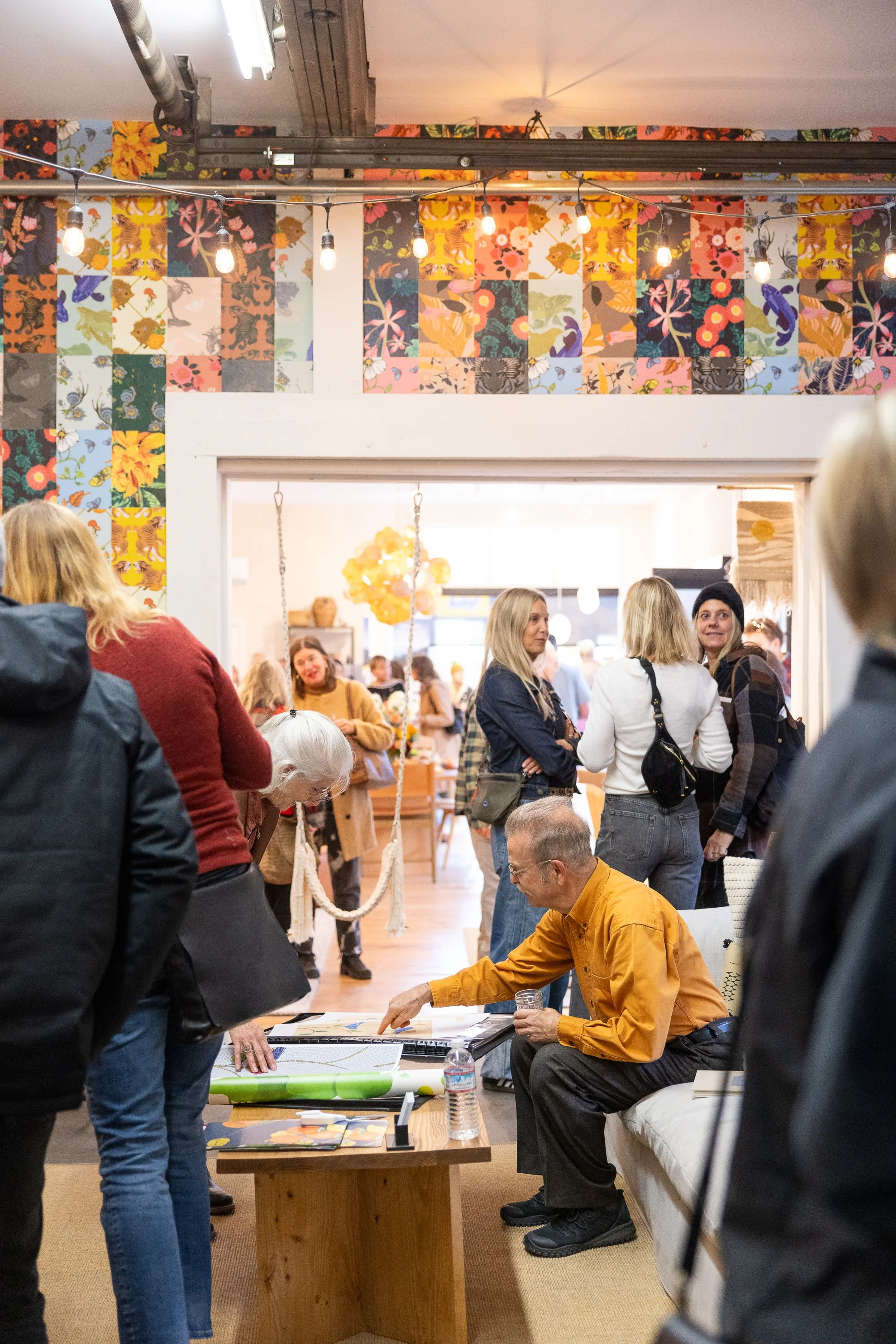 People gathered in an art or craft workshop space with colorful floral patterned wall art and string lights. Some individuals are interacting around tables, engaging with artwork or craft materials.