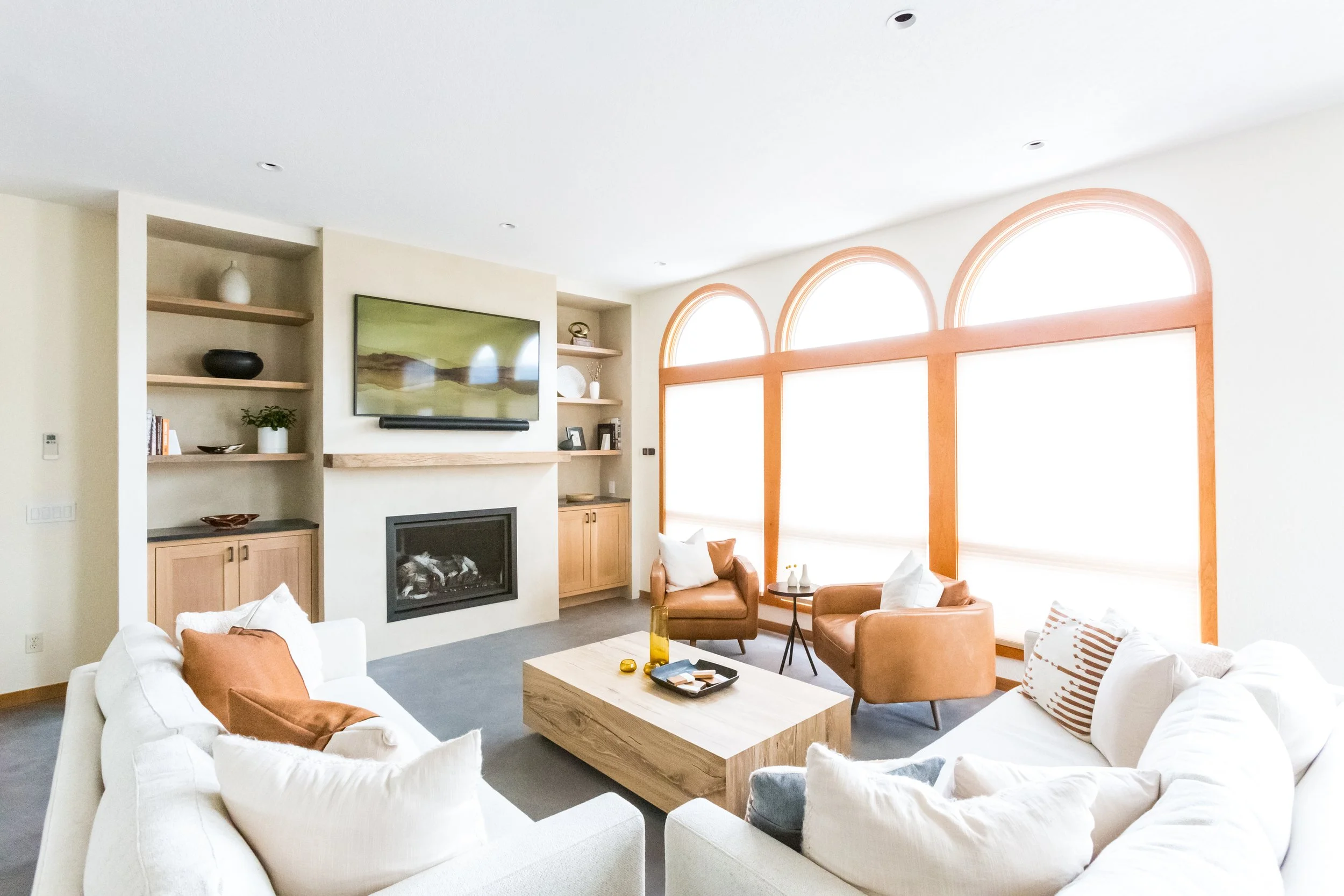 A bright living room with large arched windows, white sofas with rust-colored pillows, a wooden coffee table, brown leather armchairs, a modern fireplace, and built-in shelves with decorative items.