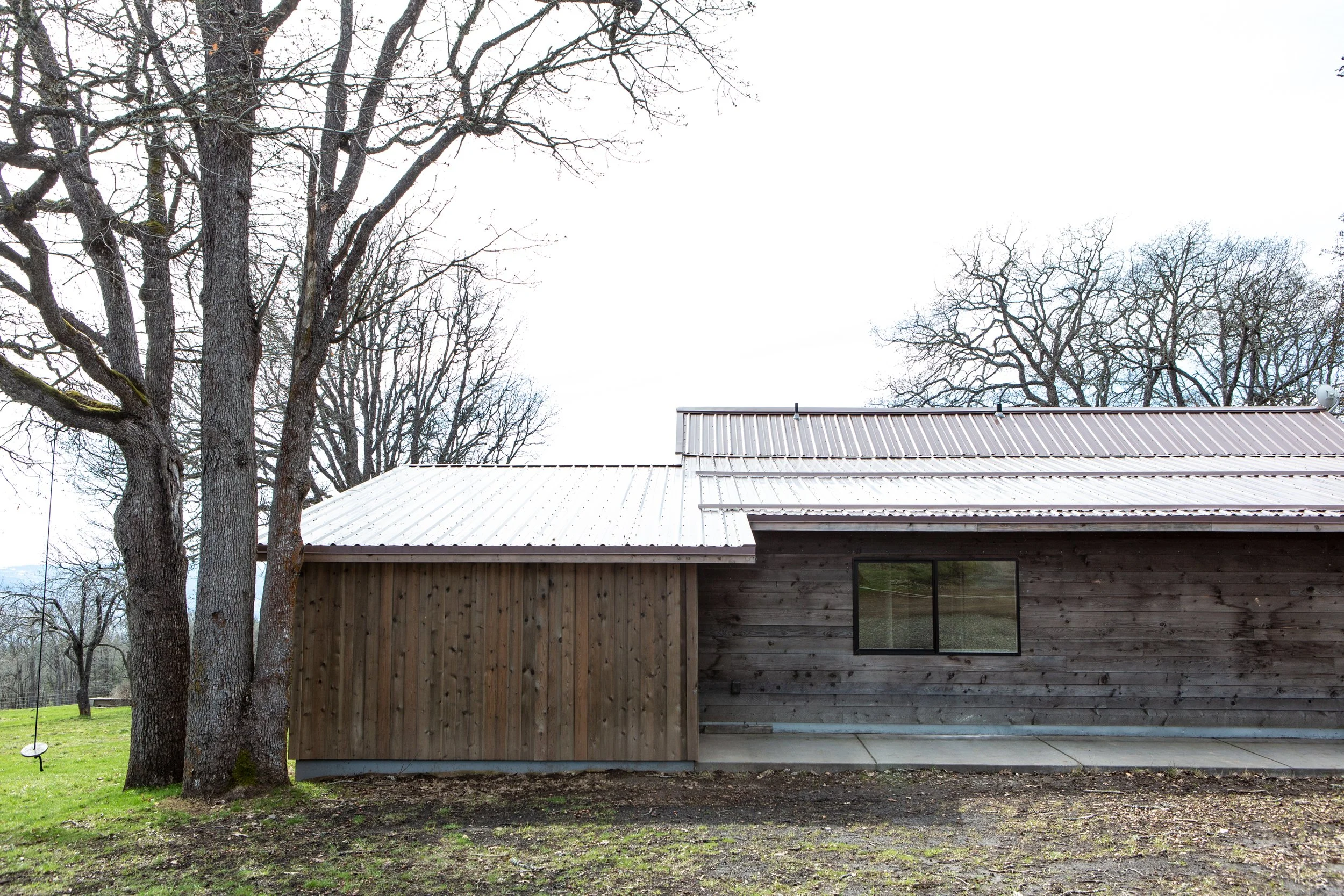 The image shows a modern wooden house with a metal roof, surrounded by leafless trees and a grassy yard.