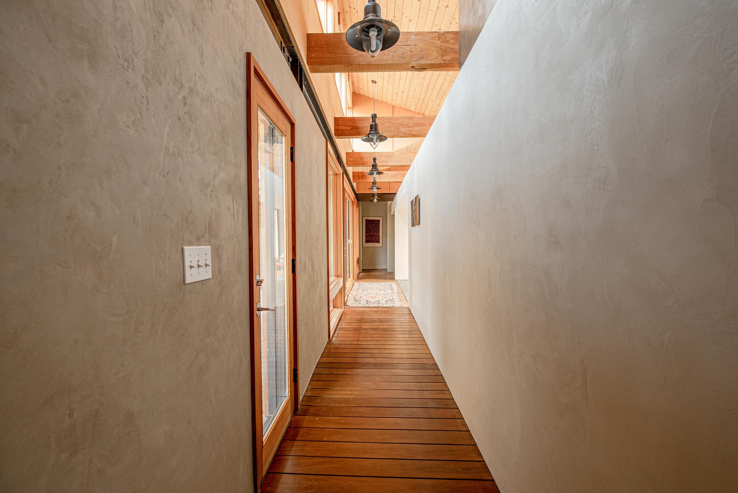 A hallway with a textured beige wall on the left, a white wall on the right, wooden doors with glass panels on the left, a wooden floor, and a ceiling with exposed wooden beams and hanging industrial-style light fixtures.