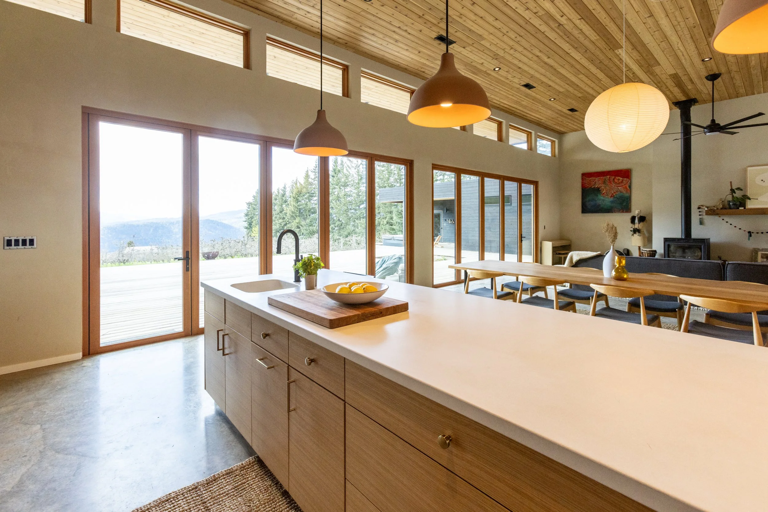 Interior of a modern home with wooden ceiling, large glass doors, a kitchen island with a white countertop, and a dining area with a wooden table and black chairs. Large windows and glass doors reveal a scenic outdoor landscape.