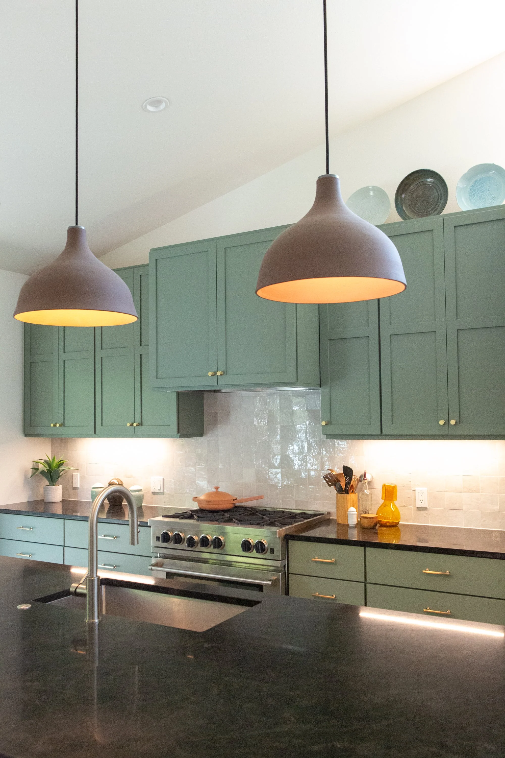 Kitchen with green cabinets, black countertop, stainless steel stove, two brown pendant lights, and various kitchen items.