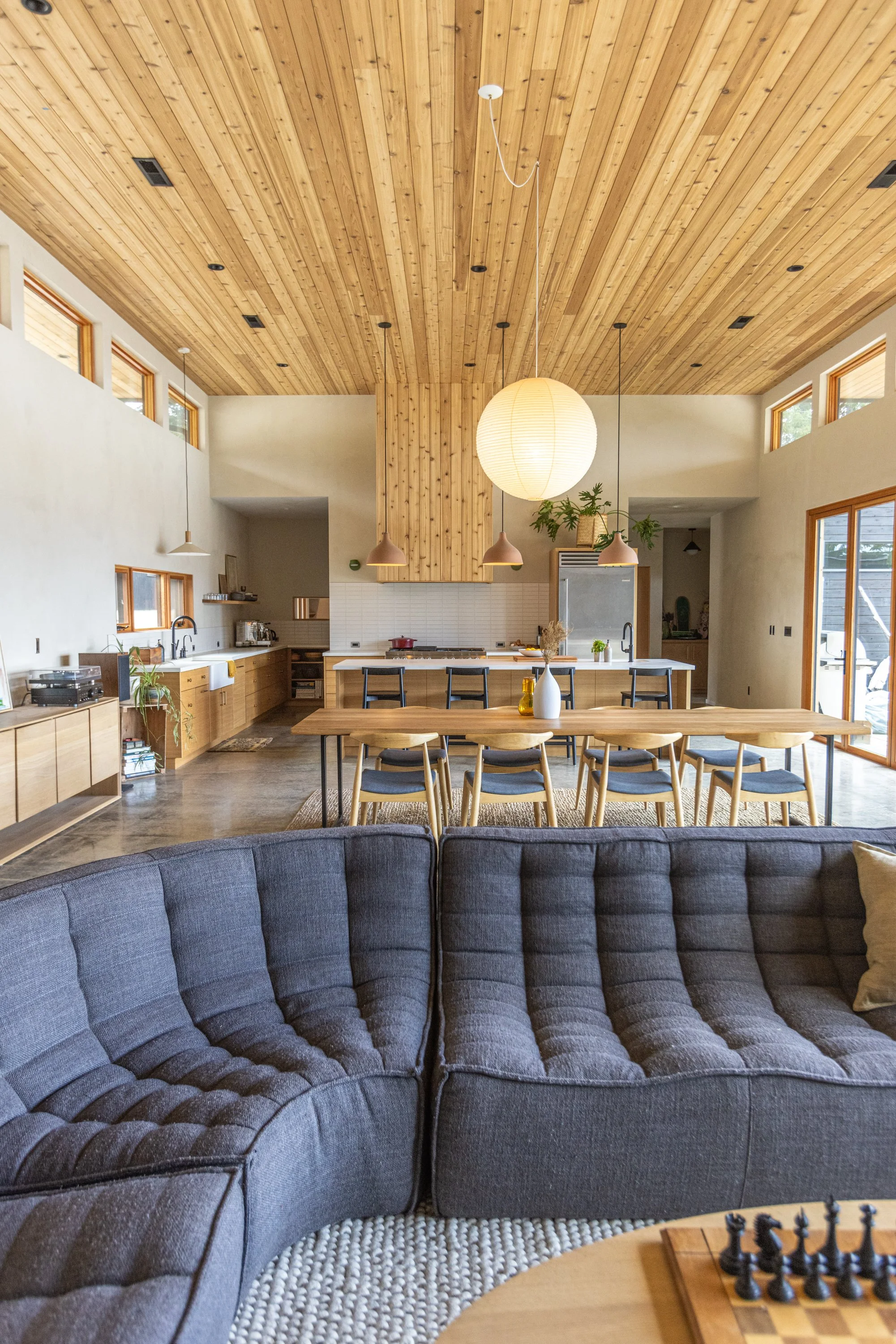 Interior of a modern kitchen and living space with a wooden ceiling, windowed walls, and a grey curved couch in the foreground. There is a dining table with chairs, a chessboard, and a kitchen counter with appliances in the background.