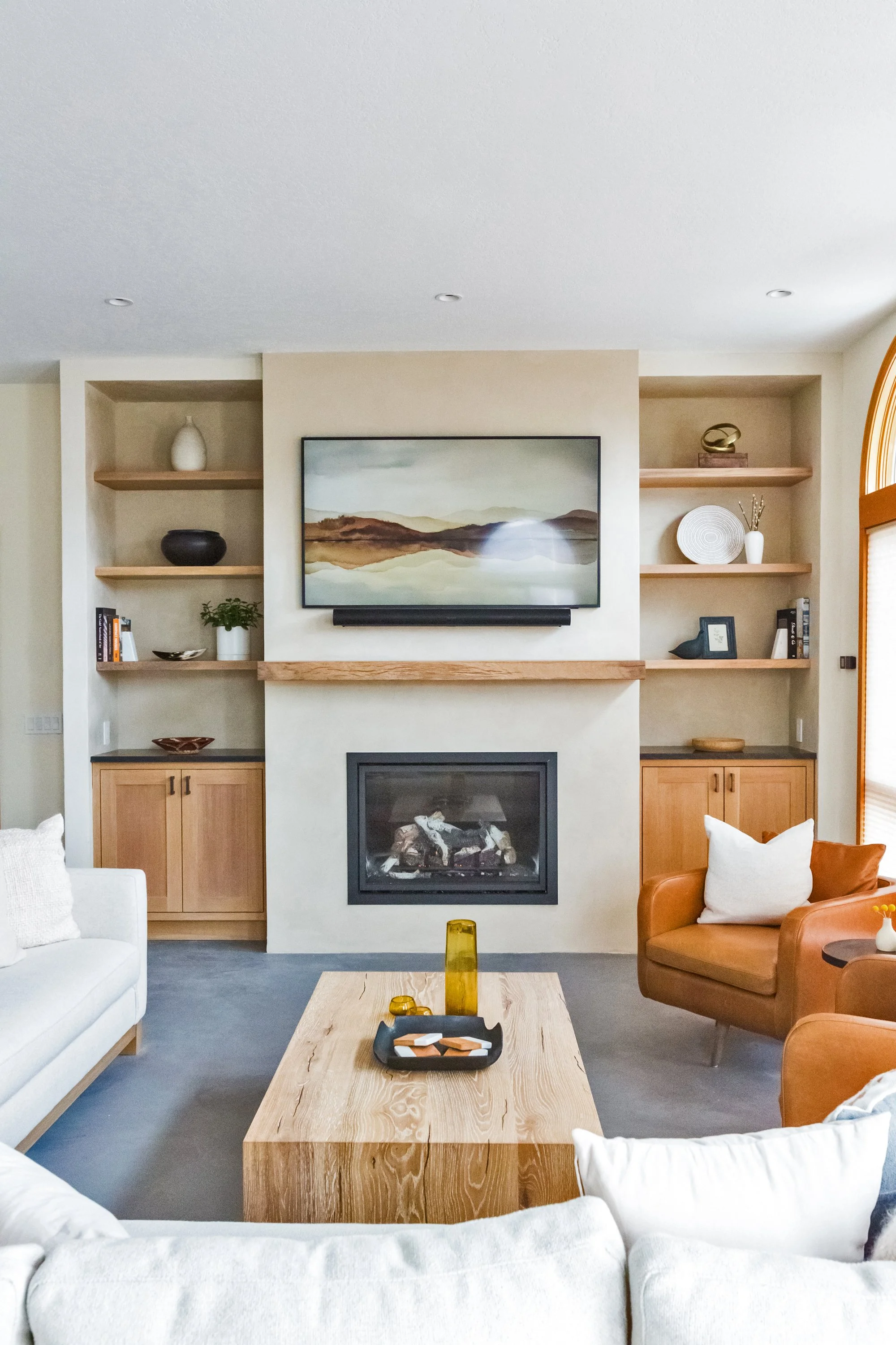 Living room with a fireplace, wall-mounted TV, built-in shelves with decorative items, a white sofa, and an orange armchair, with a wooden coffee table and yellow candle holders.