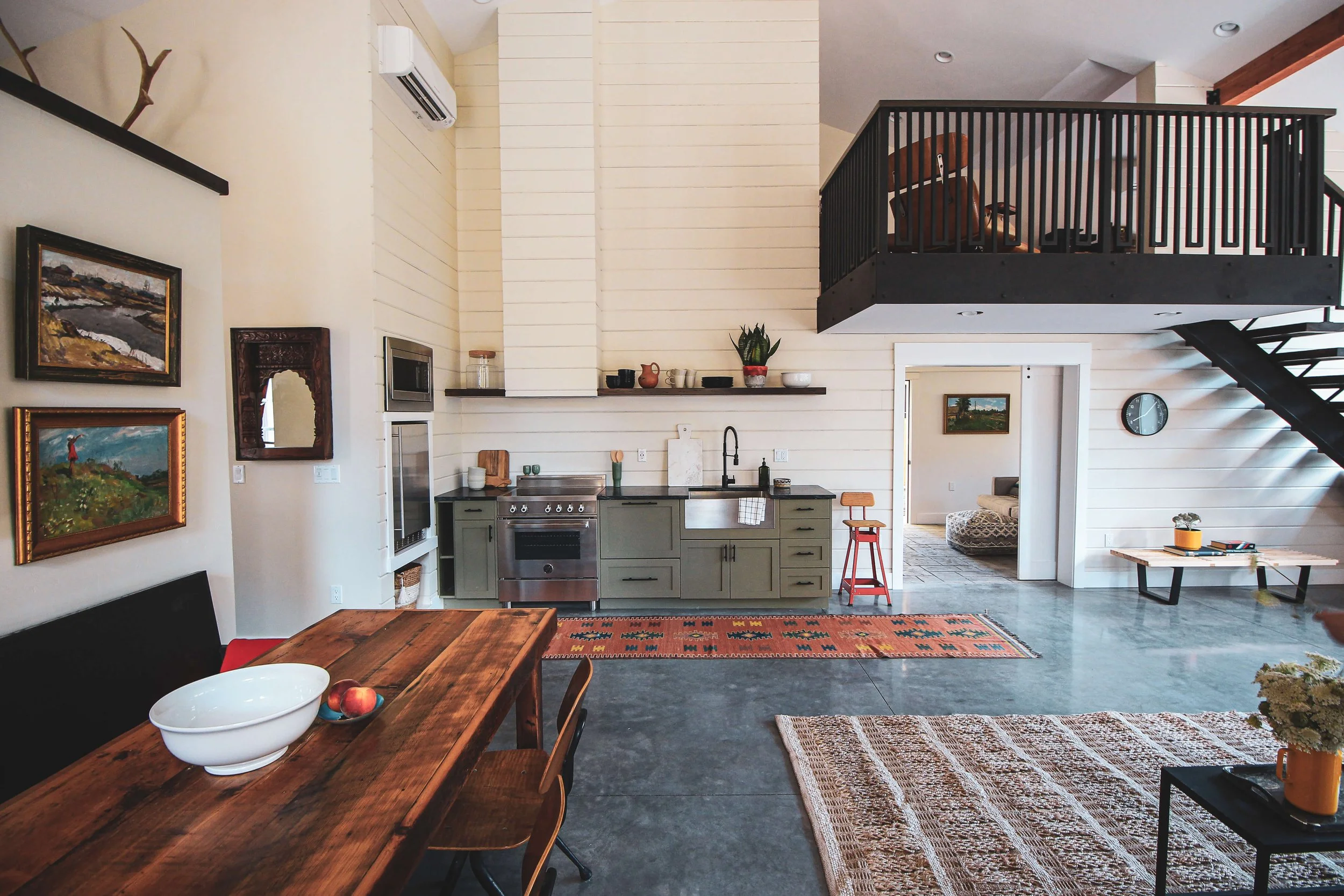 A spacious, modern kitchen and living area with a high ceiling and white shiplap walls. Features a dark dining table with wooden chairs, green kitchen cabinets, stainless steel appliances, and open shelving with dishes. A black staircase leads to an 