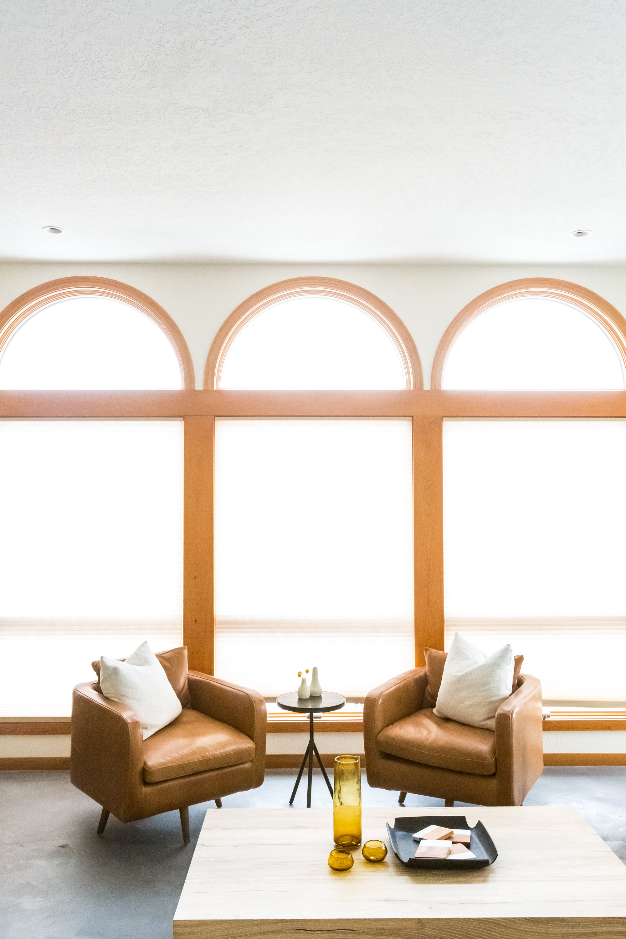 Living room with two brown leather armchairs, a small round side table, and three yellow glass vases, set against large arched windows with wooden trim.
