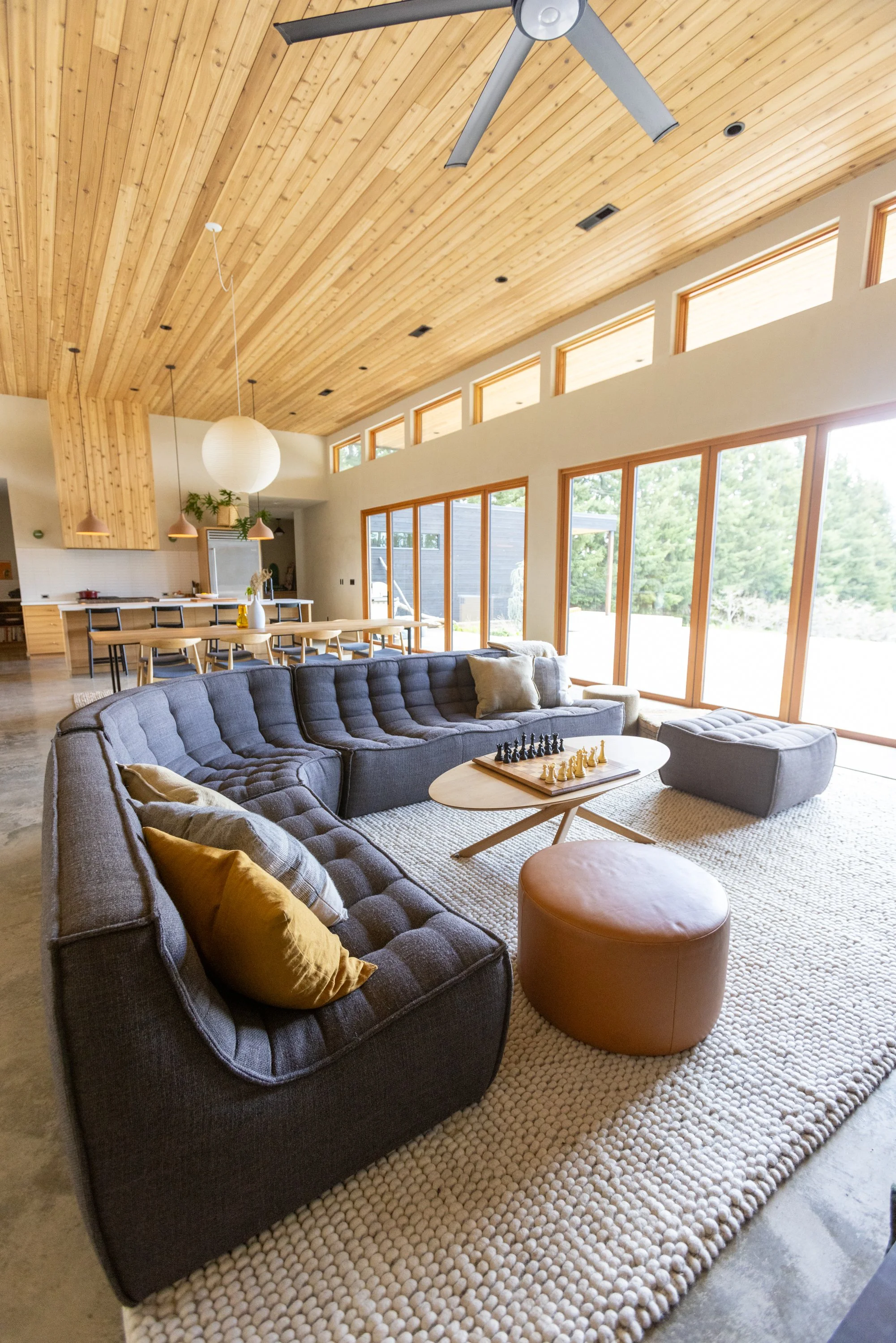 Living room with large gray couch, tan pillows, round brown ottoman, and chessboard on a white rug, high wood ceiling with skylights, large windows, and dining area with wooden table and chairs.