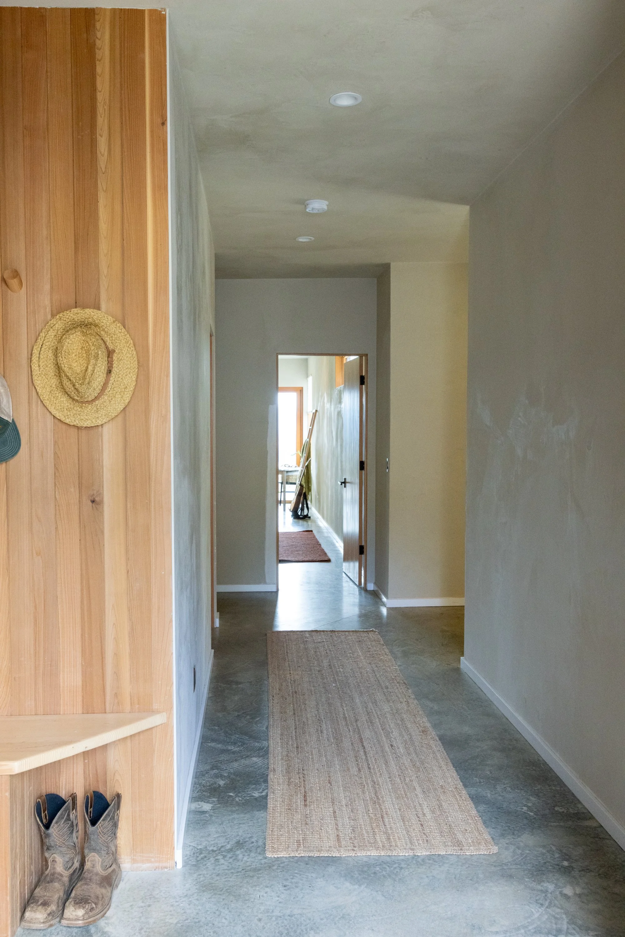 A hallway with wooden steps on the left, cowboy boots at the bottom of the steps, and a hallway with a rug leading to a room with a window and a table.