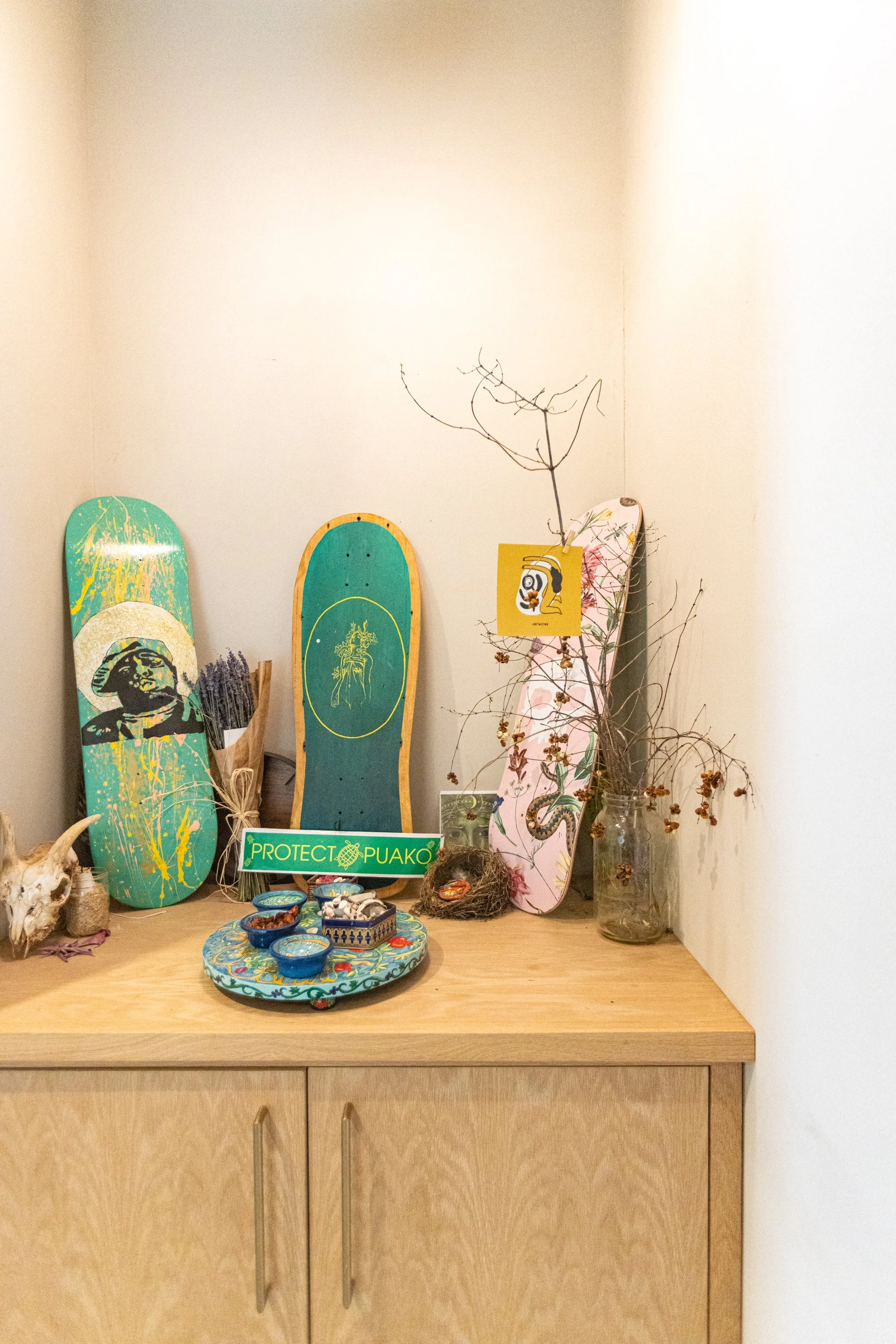 Display of decorative skateboards, plants, and bowls on a wooden table with a beige cabinet below. A sign reads 'Protect Puako'.