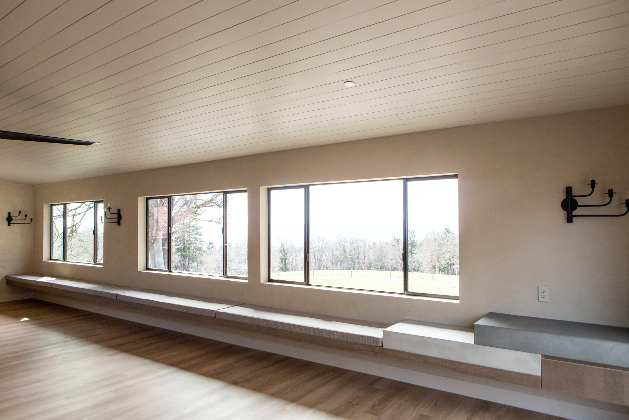 Interior of a room with large windows, hardwood flooring, a white ceiling with wood paneling, black wall sconces, and a grey bench under the windows.