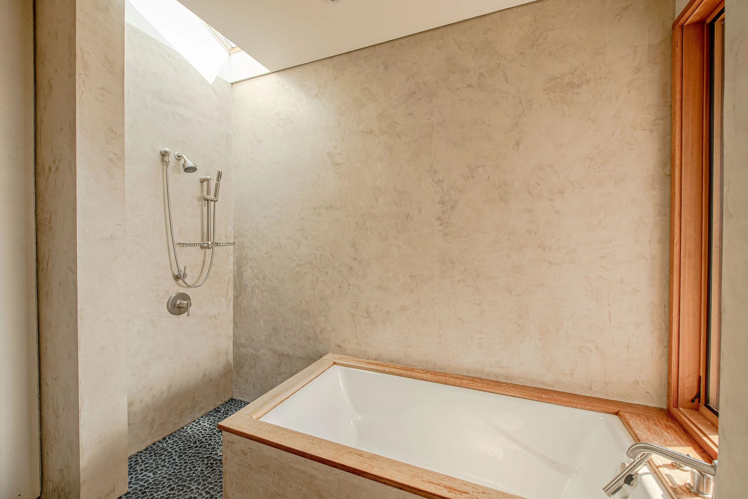 Bathroom with a shower area and a bathtub, featuring beige textured walls, a skylight, a showerhead, and a wooden-framed mirror.