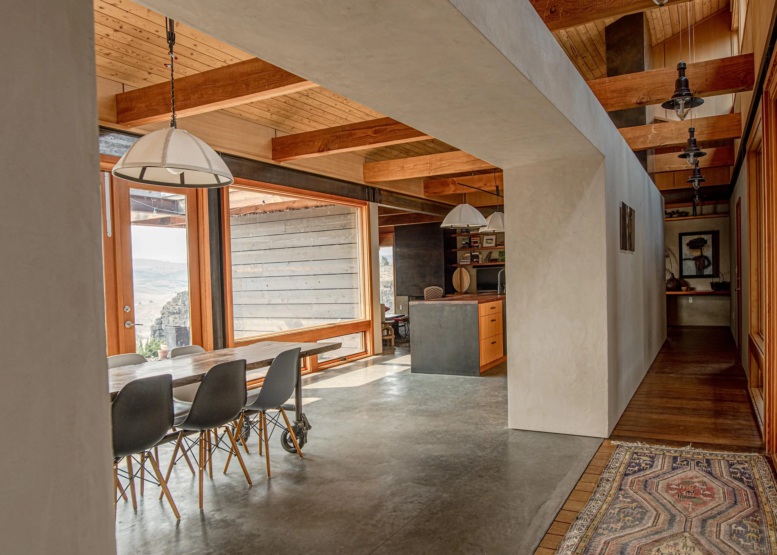 Interior view of a modern house with wooden ceilings, large glass windows, a dining table with black chairs, and decorative lighting fixtures.