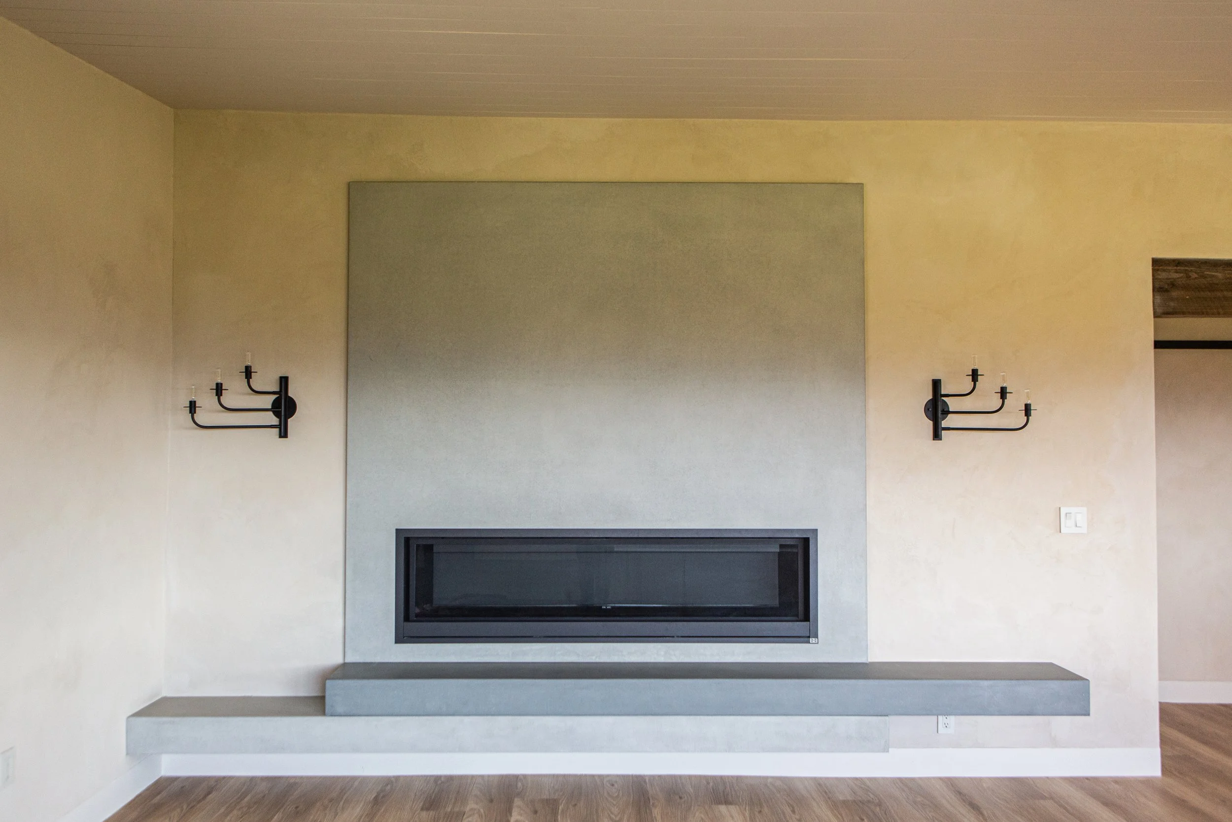 Living room with a gray wall, fireplace, and black wall sconces on each side of the fireplace, with a wooden floor.