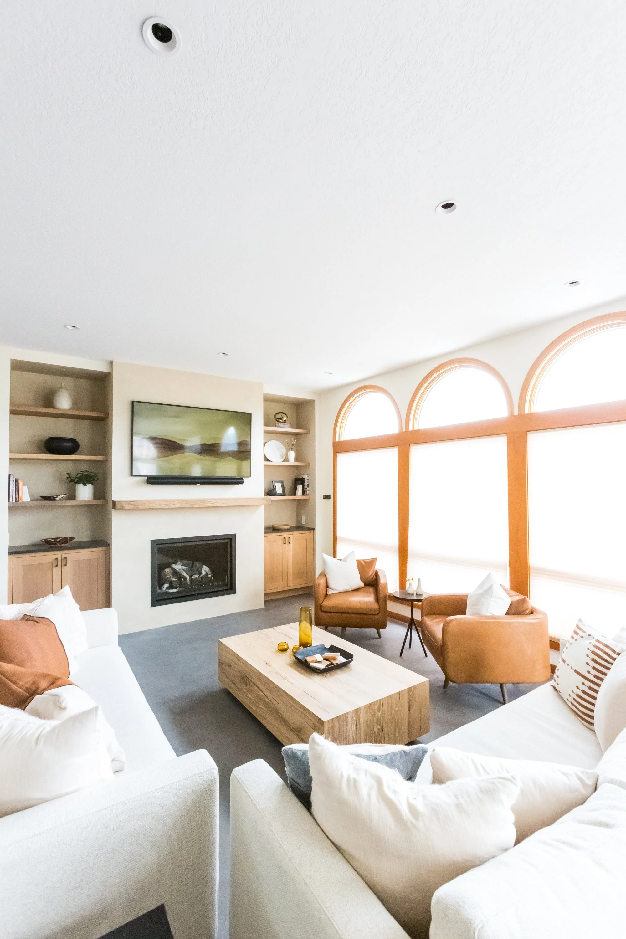 A living room with large windows, a fireplace, television, beige built-in shelves, and brown leather chairs arranged around a light-colored wooden coffee table. White cushions are on the sofas, and decorative objects and books are on the shelves.
