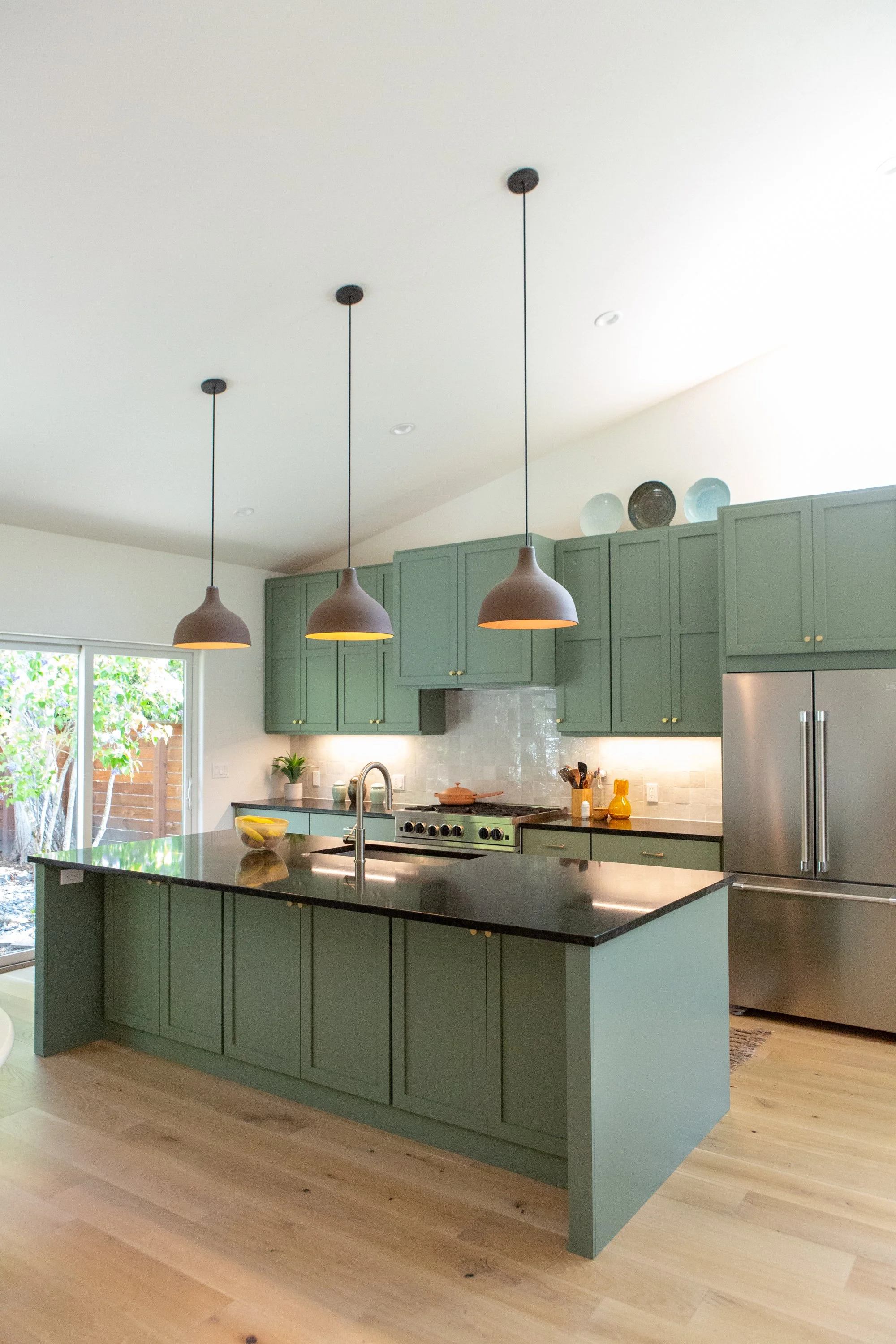 A modern kitchen with green cabinets, black countertop island, stainless steel refrigerator, and three pendant lights hanging from the ceiling.