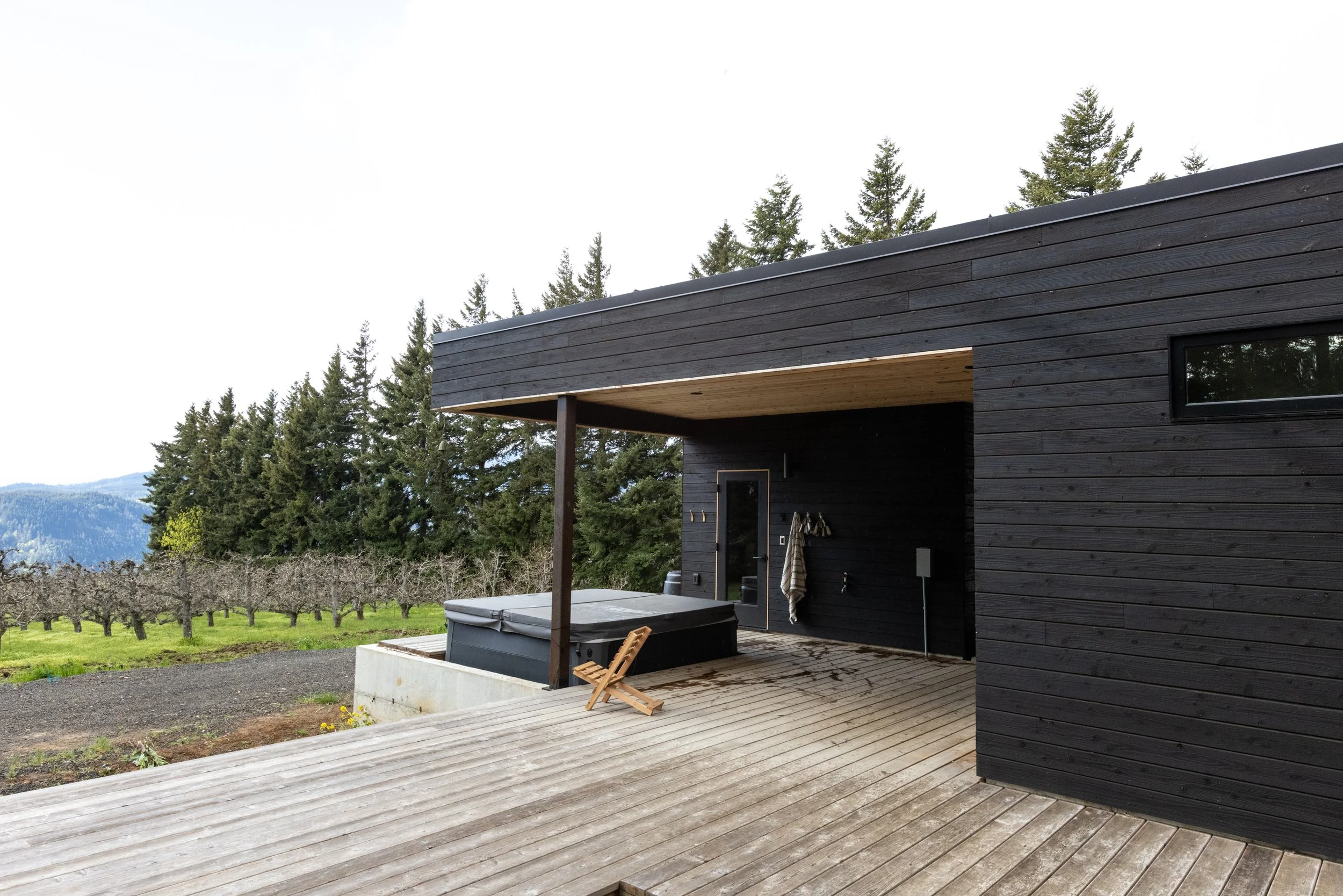 Modern black wooden house with a deck, hot tub, and outdoor furniture, surrounded by trees and mountains in the background.