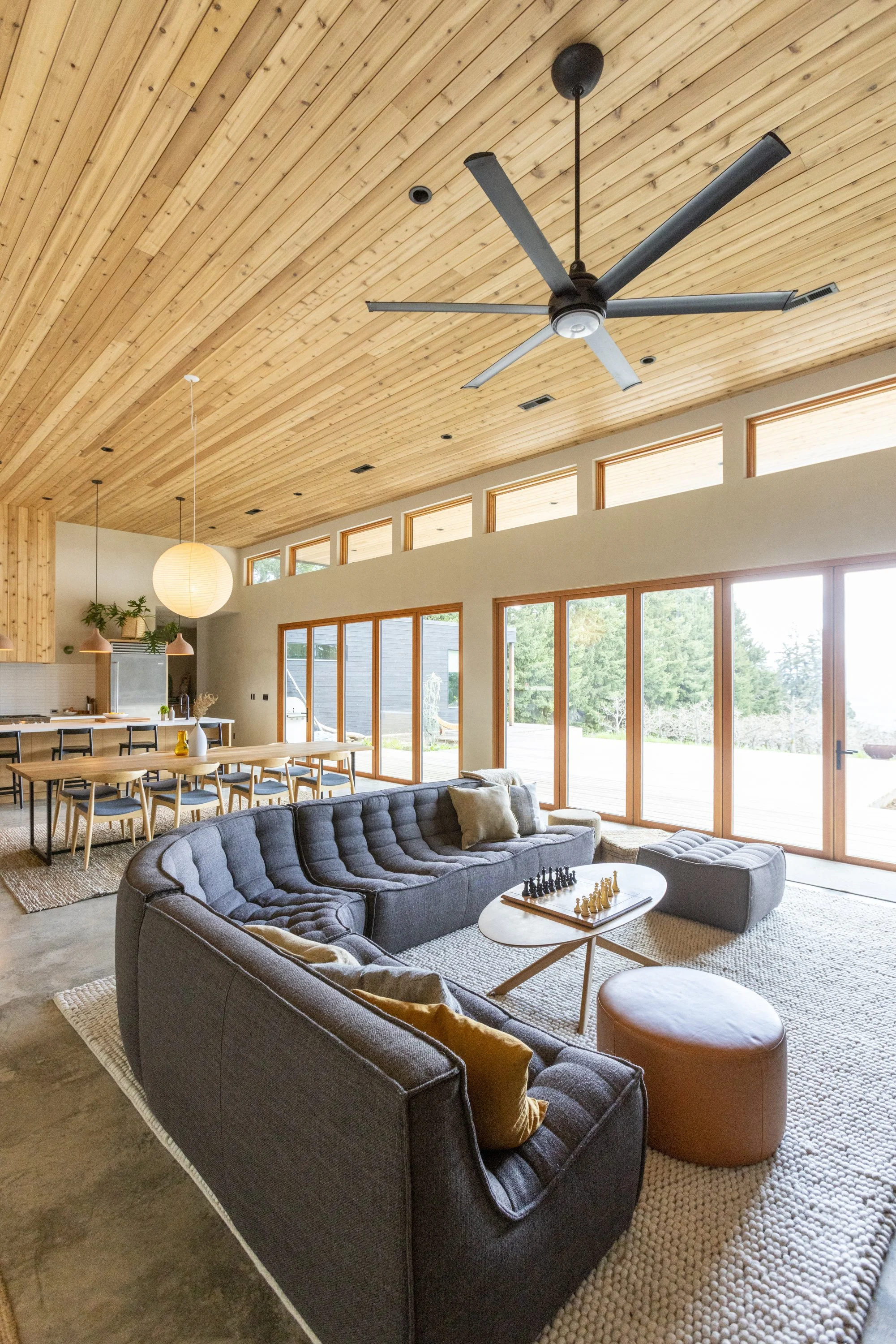 Living room with a dark gray curved sofa, a small round white coffee table with a chessboard, a beige pouf, a beige textured rug, large glass doors letting in natural light, and a wood ceiling with a ceiling fan.