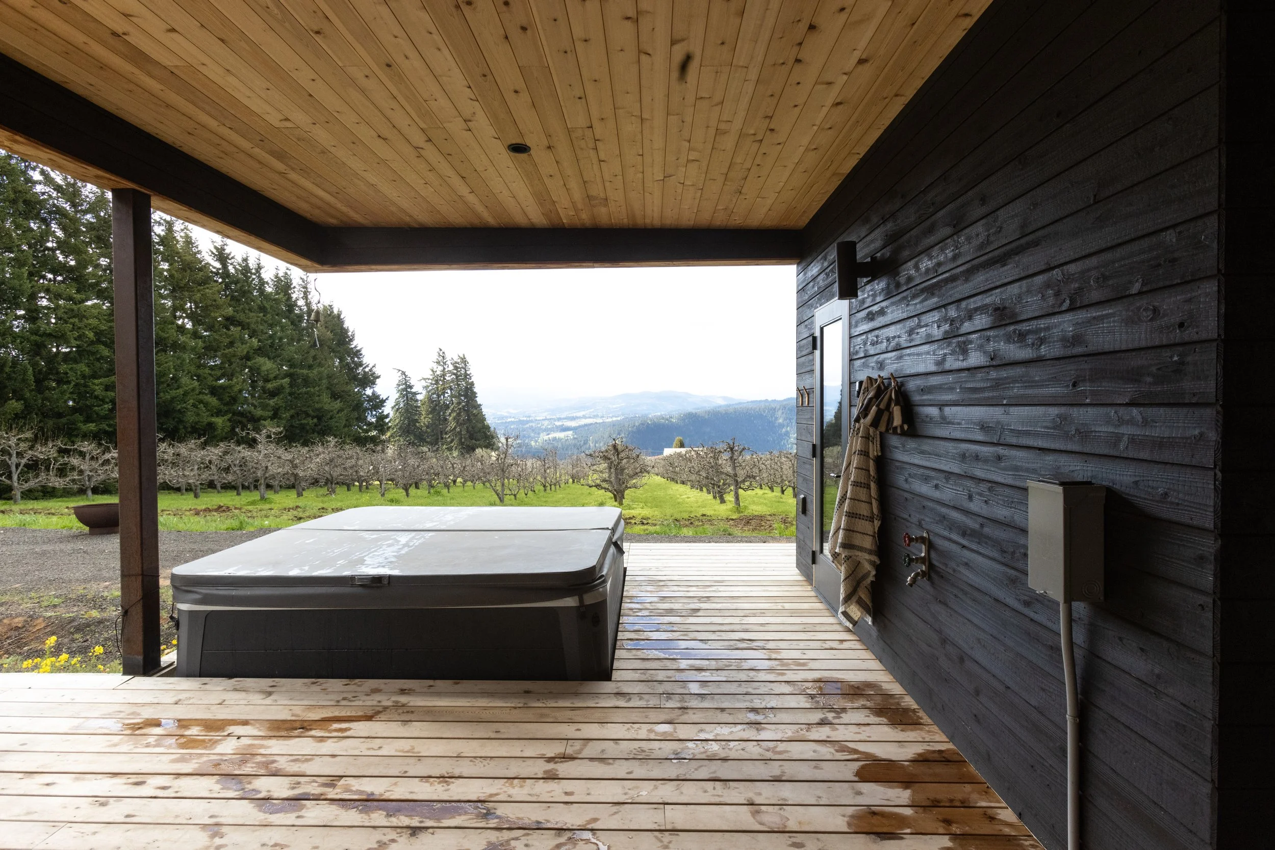 View from a covered patio showing a hot tub, a wooden deck, a dark wooden wall with electric meter, a towel hanging, and an outdoor landscape with trees and distant hills.