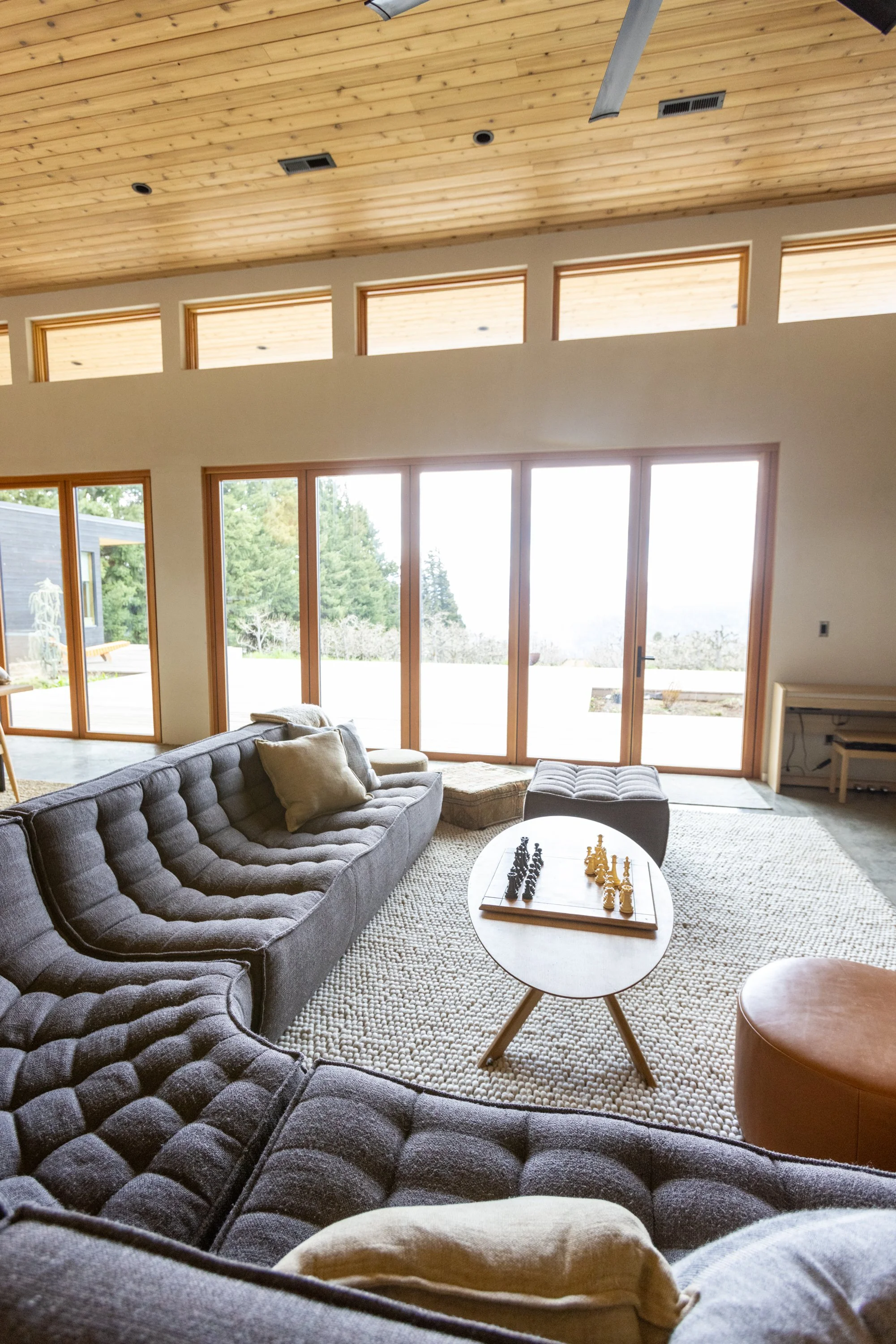 Living room with large windows, gray tufted sofa with pillows, round coffee table with chess set, beige carpet, wooden ceiling, and natural light.