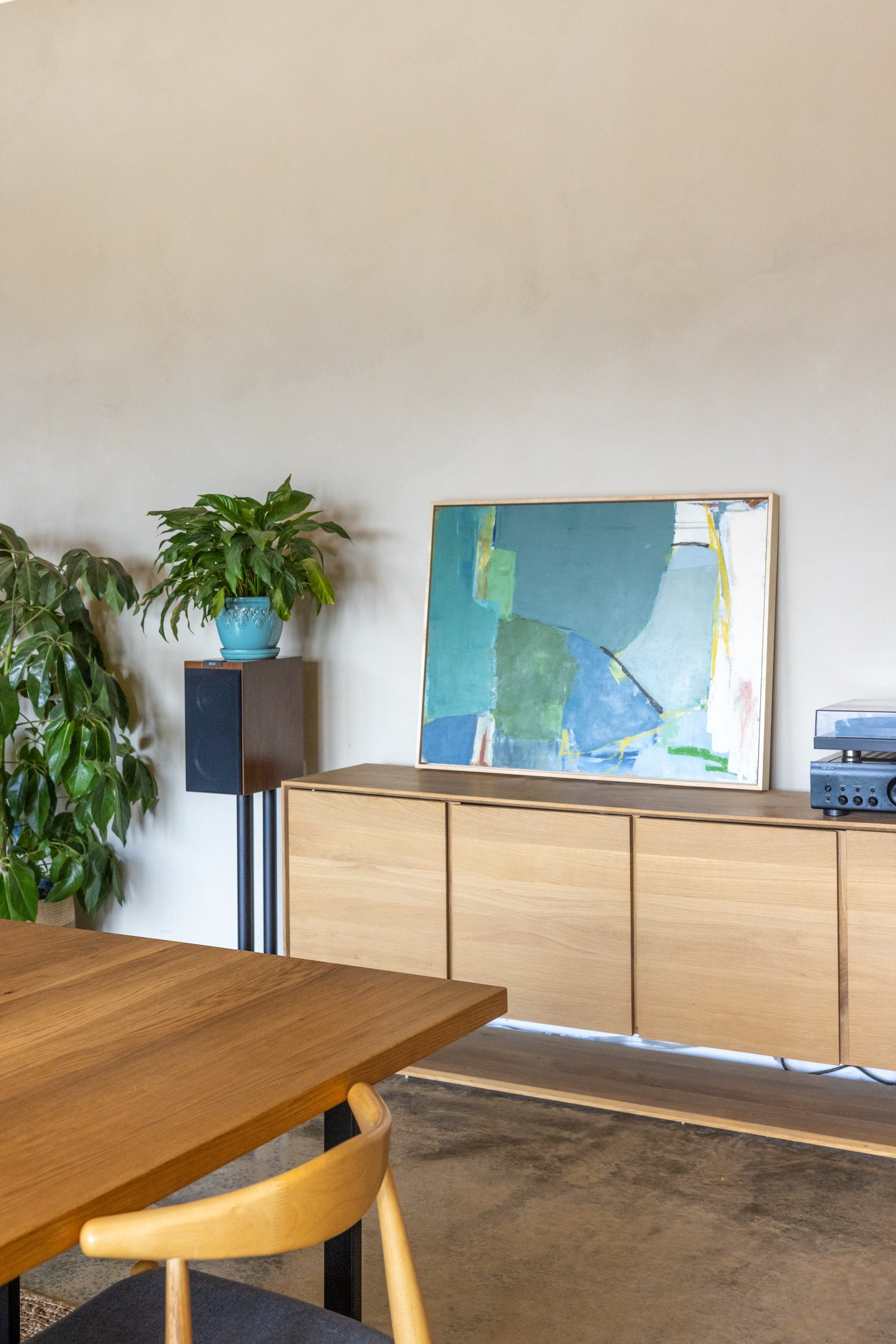 Interior of a modern, minimalist room with a wooden table, beige wall, plants, and abstract artwork on a wooden sideboard.