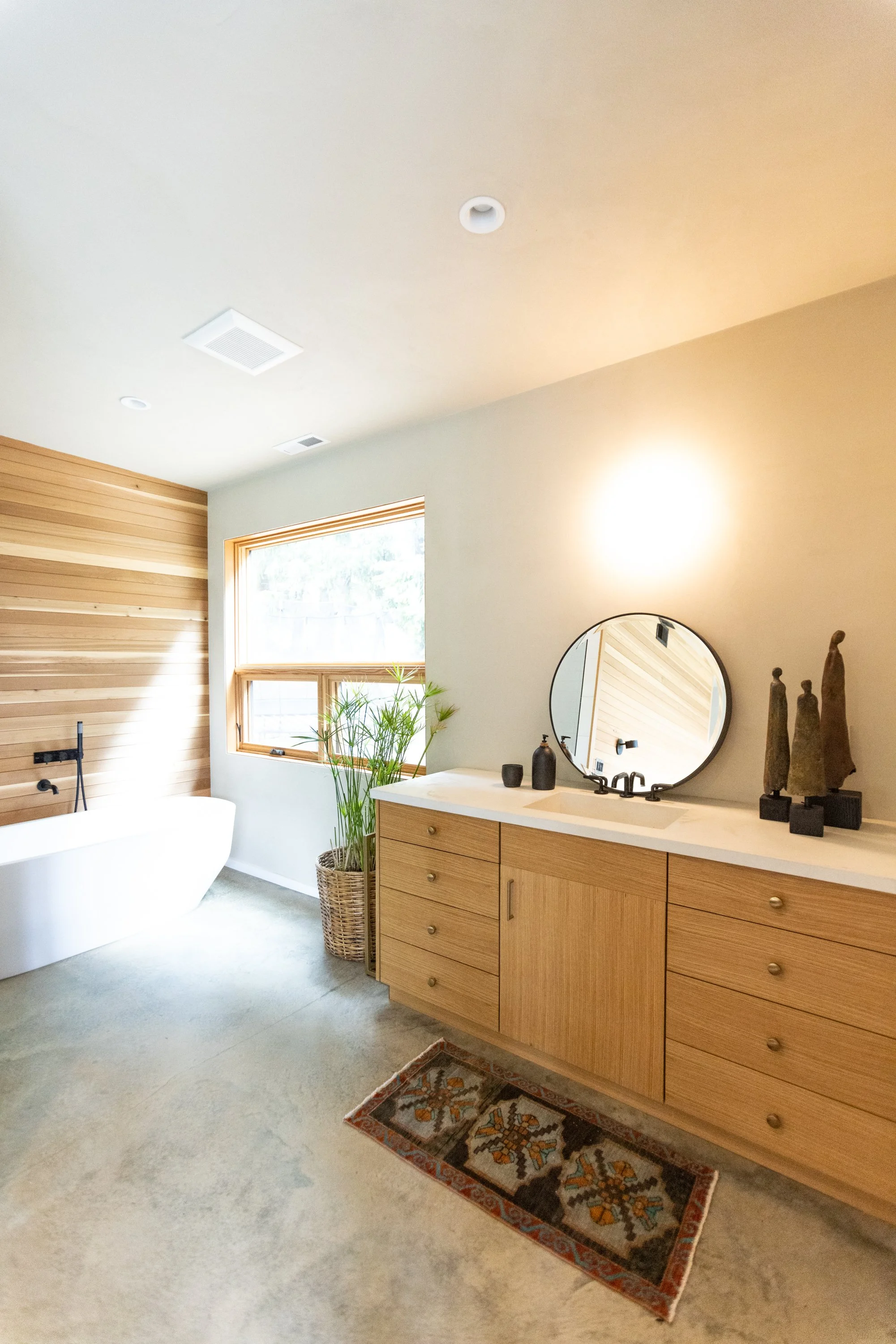 A bathroom with a freestanding bathtub, wooden vanity with a round mirror, and a window letting in natural light, decorated with indoor plants and sculptures.