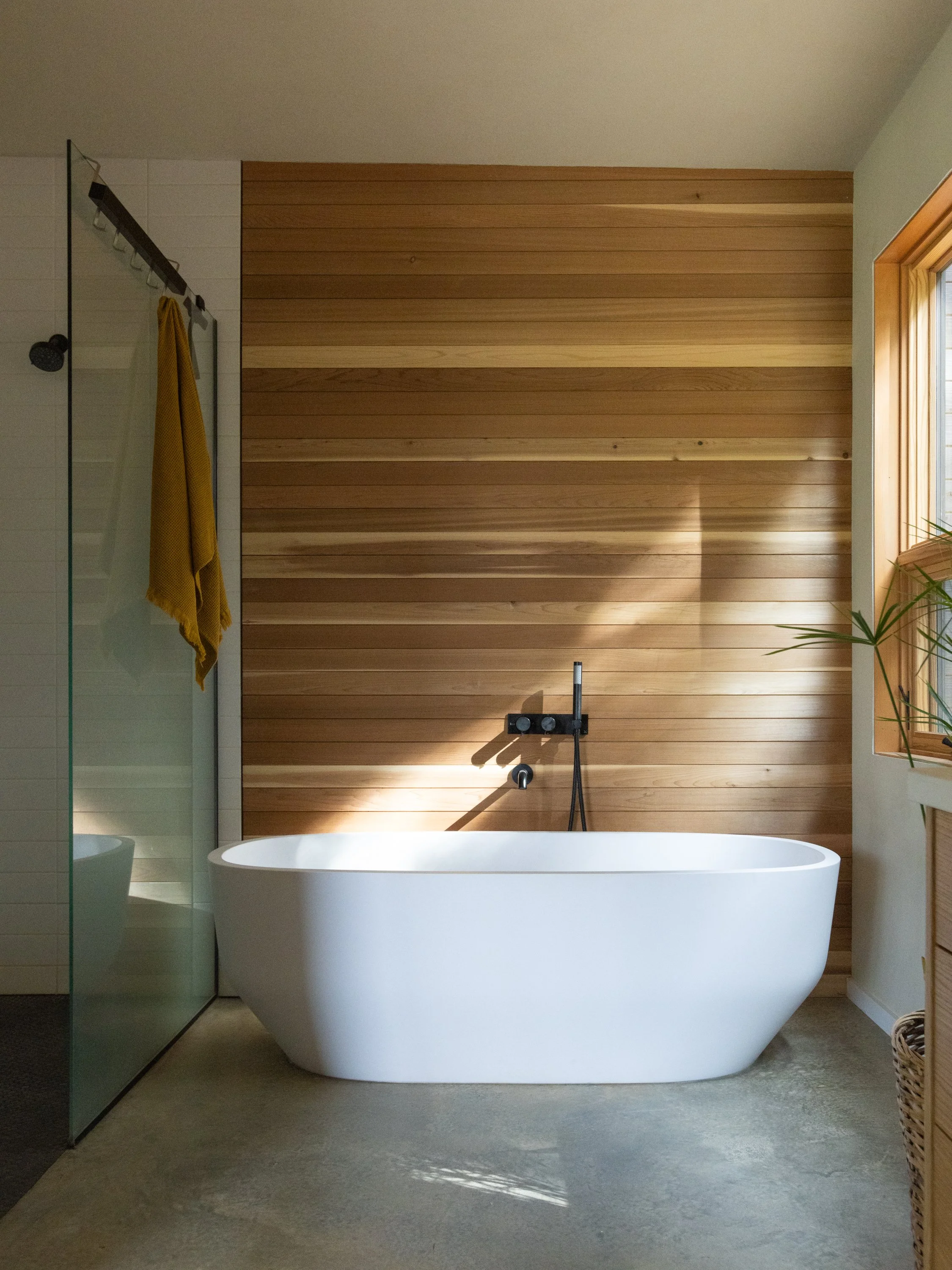 Modern bathroom with a white freestanding bathtub, wood-paneled accent wall, large window, and concrete floor.
