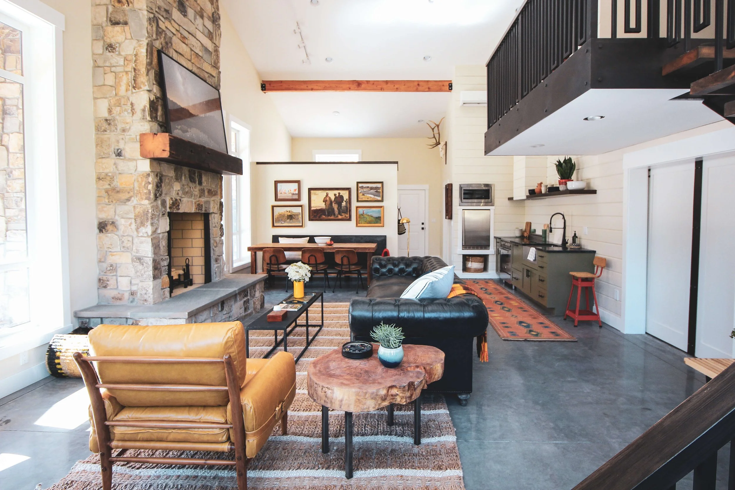 A cozy living room with a stone fireplace, a wooden dining table, and a modern kitchen area. The space features a blend of rustic and contemporary decor, with artwork on the walls, a leather sofa, and wooden furniture.