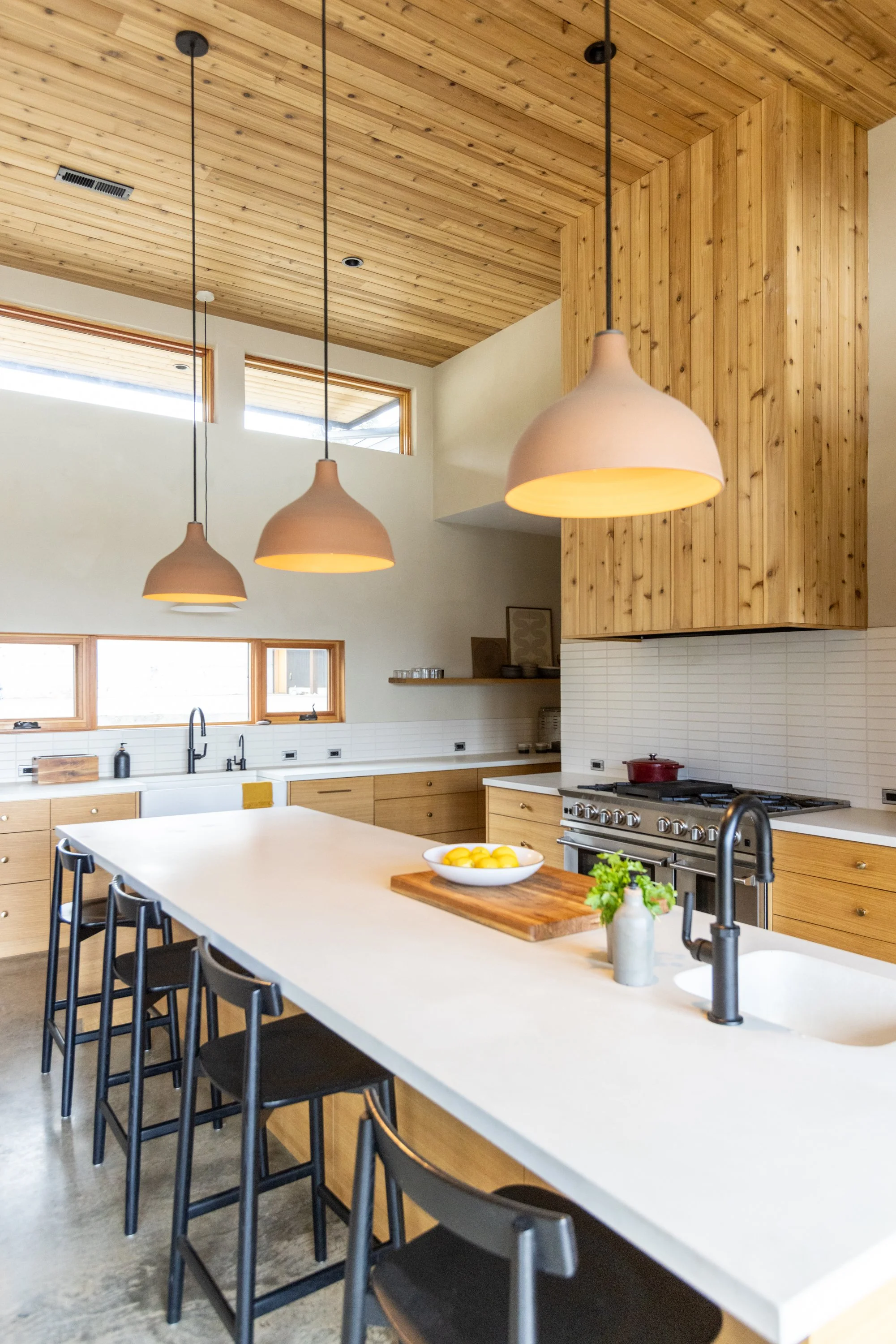 Modern kitchen with wooden upper cabinets, white countertop and backsplash, black chairs around a kitchen island, pendant lighting, and windows letting in natural light.
