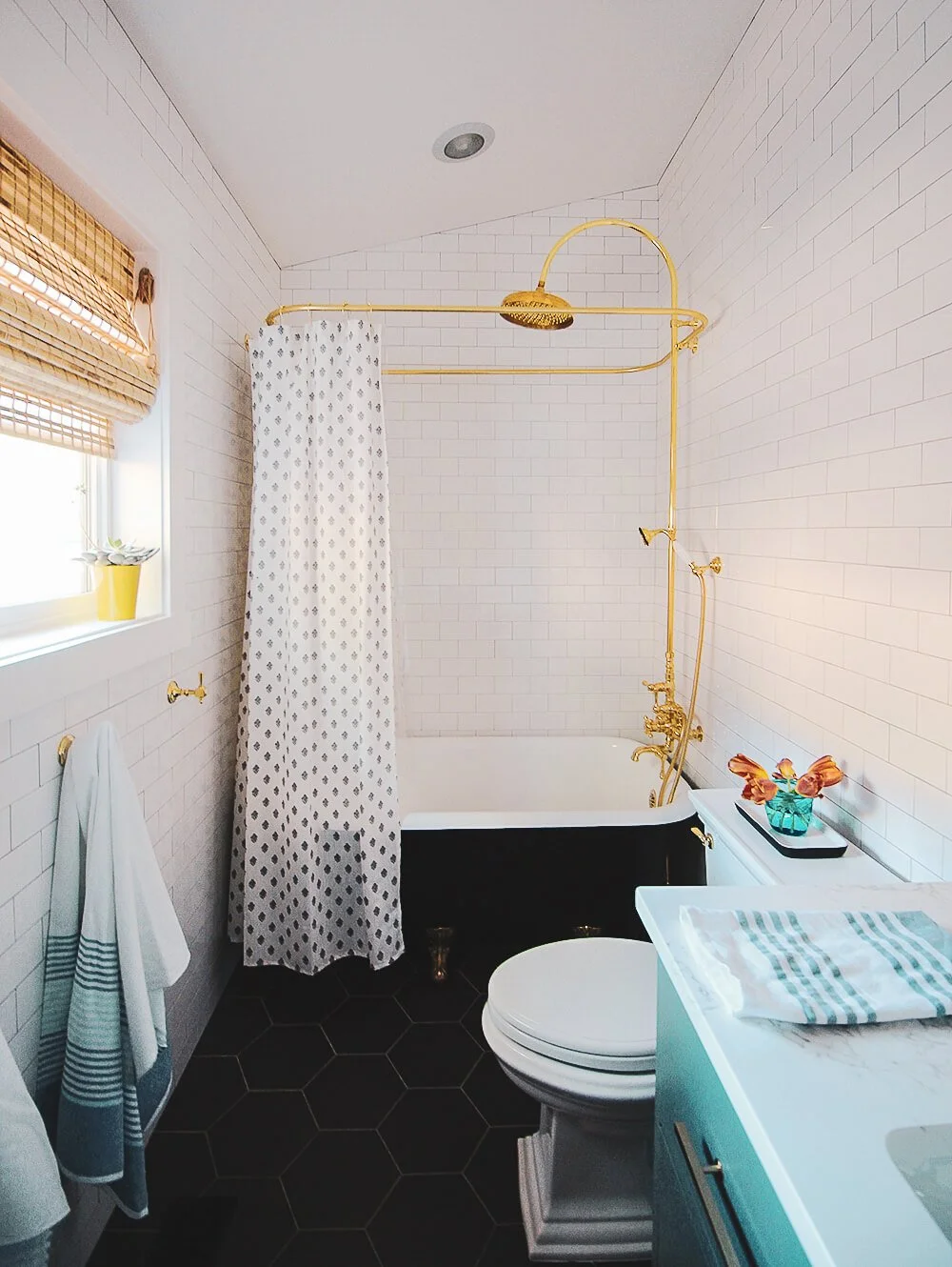 Bathroom with white brick walls, black hexagonal floor tiles, a white bathtub with black exterior and gold fixtures, a shower with gold pipe and showerhead, a small window with bamboo shades, a white vanity with a striped towel, a small vase with ora