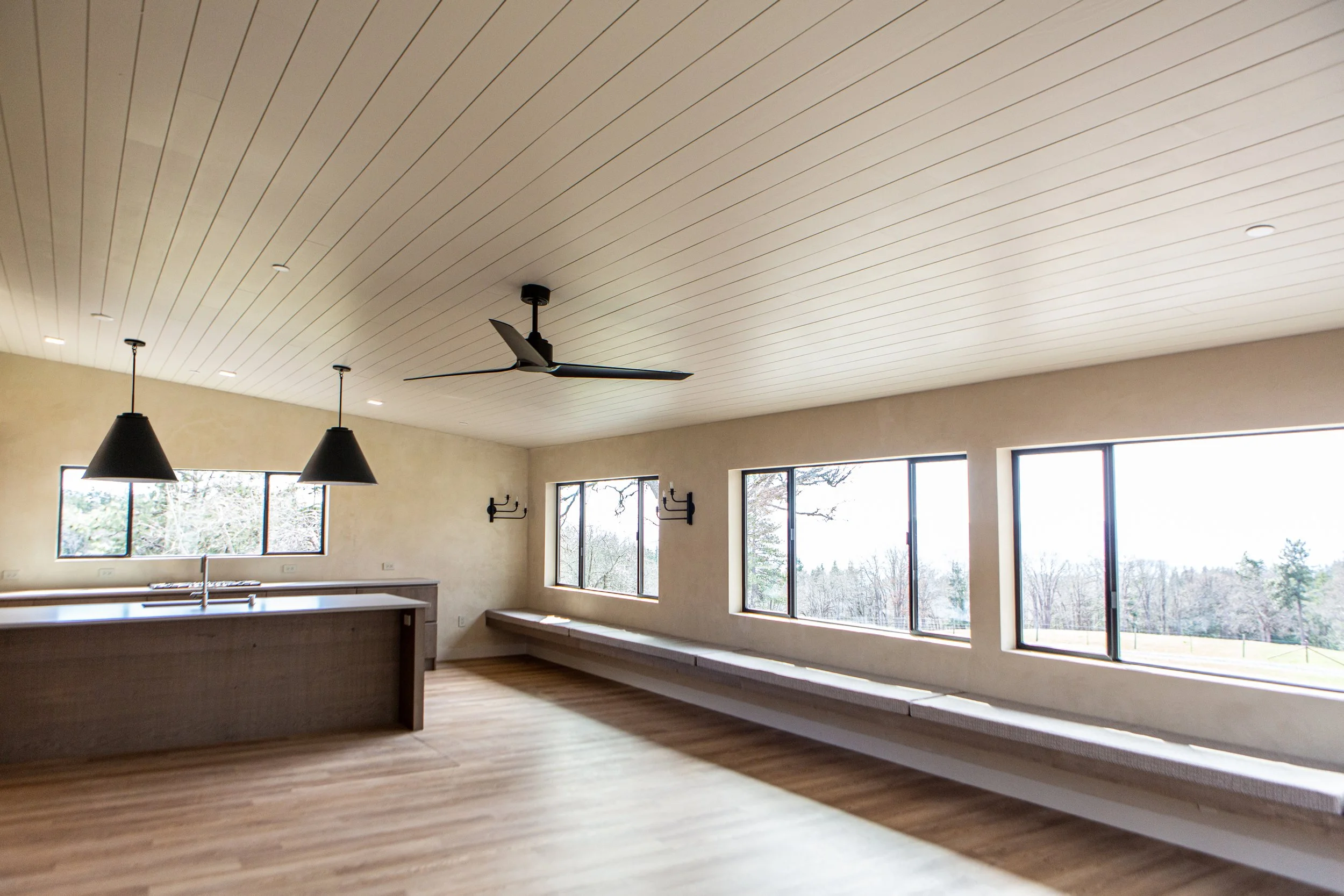 Interior view of a modern room with large windows, wooden floors, a black ceiling fan, pendant lights, and a built-in window seat under the windows.