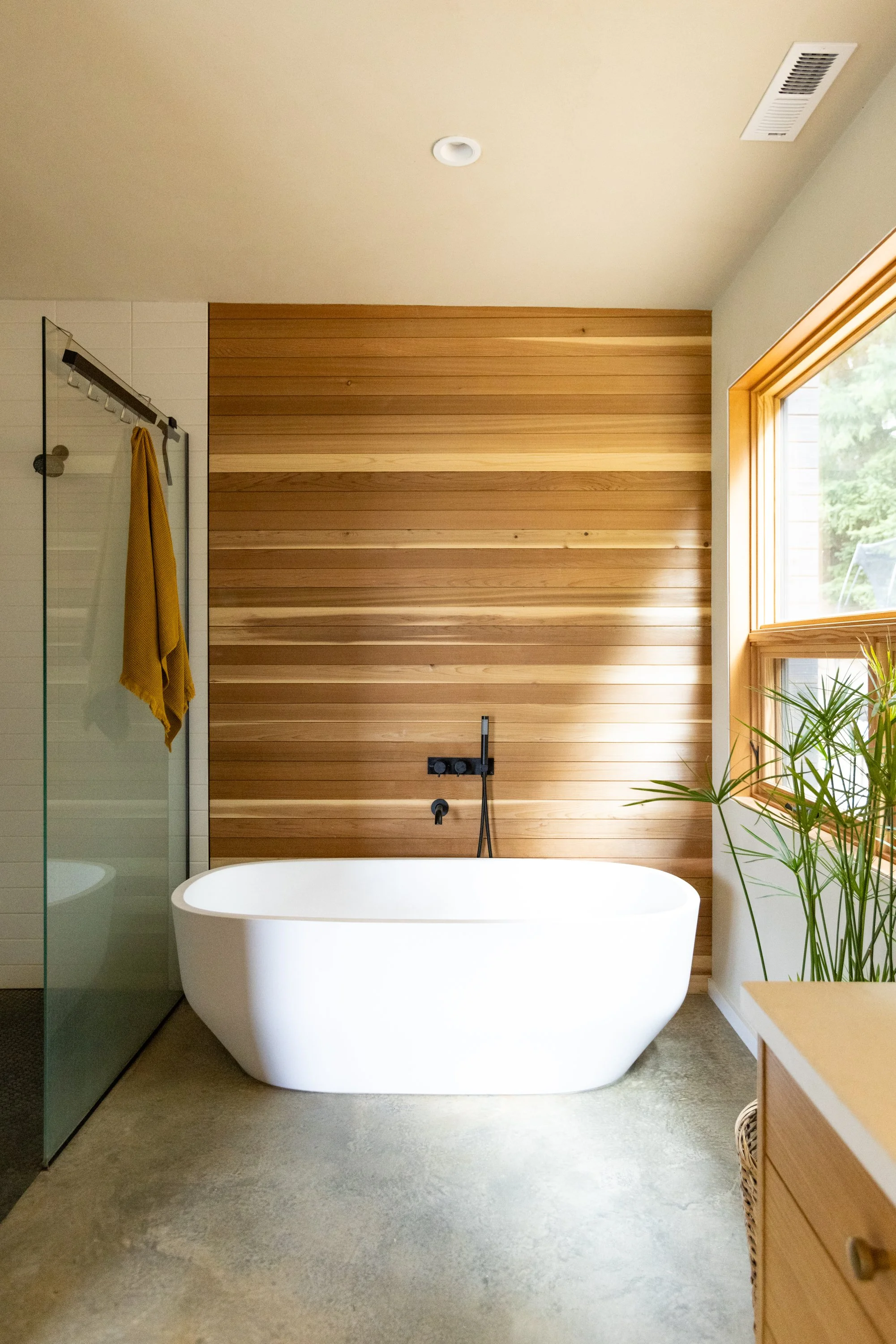 A modern bathroom featuring a white freestanding bathtub, a wooden accent wall, a large window with natural light, green plants, a glass shower enclosure with a yellow towel, and a ceiling vent.