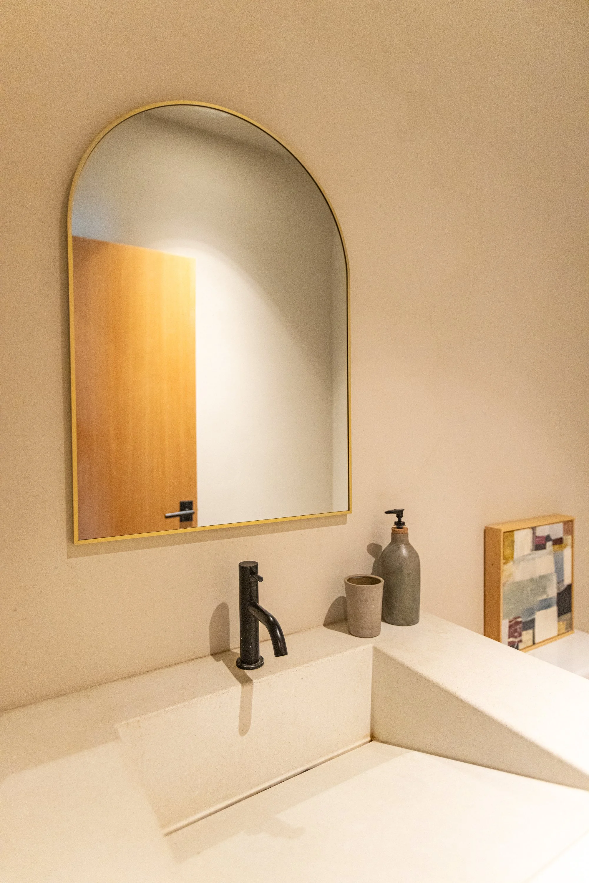A bathroom sink area with a black faucet, two decorative bottles, and a beige countertop. A mirror with a gold frame hangs above the sink, reflecting a closed wooden door. There is a small framed artwork on the wall to the right of the mirror.