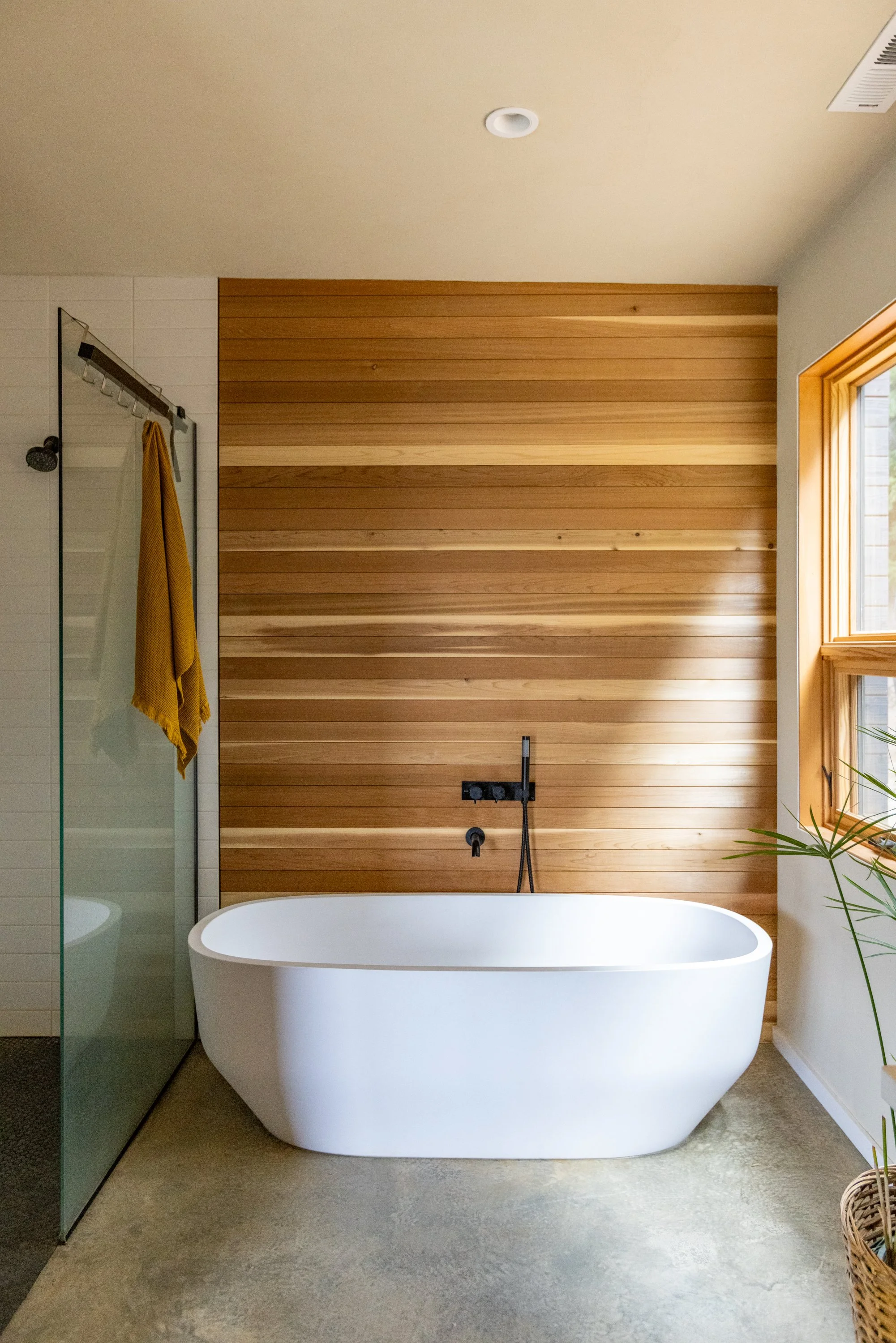 Modern bathroom with a white freestanding bathtub, wooden accent wall, window with wooden trim, and a walk-in shower with a yellow towel hanging inside.