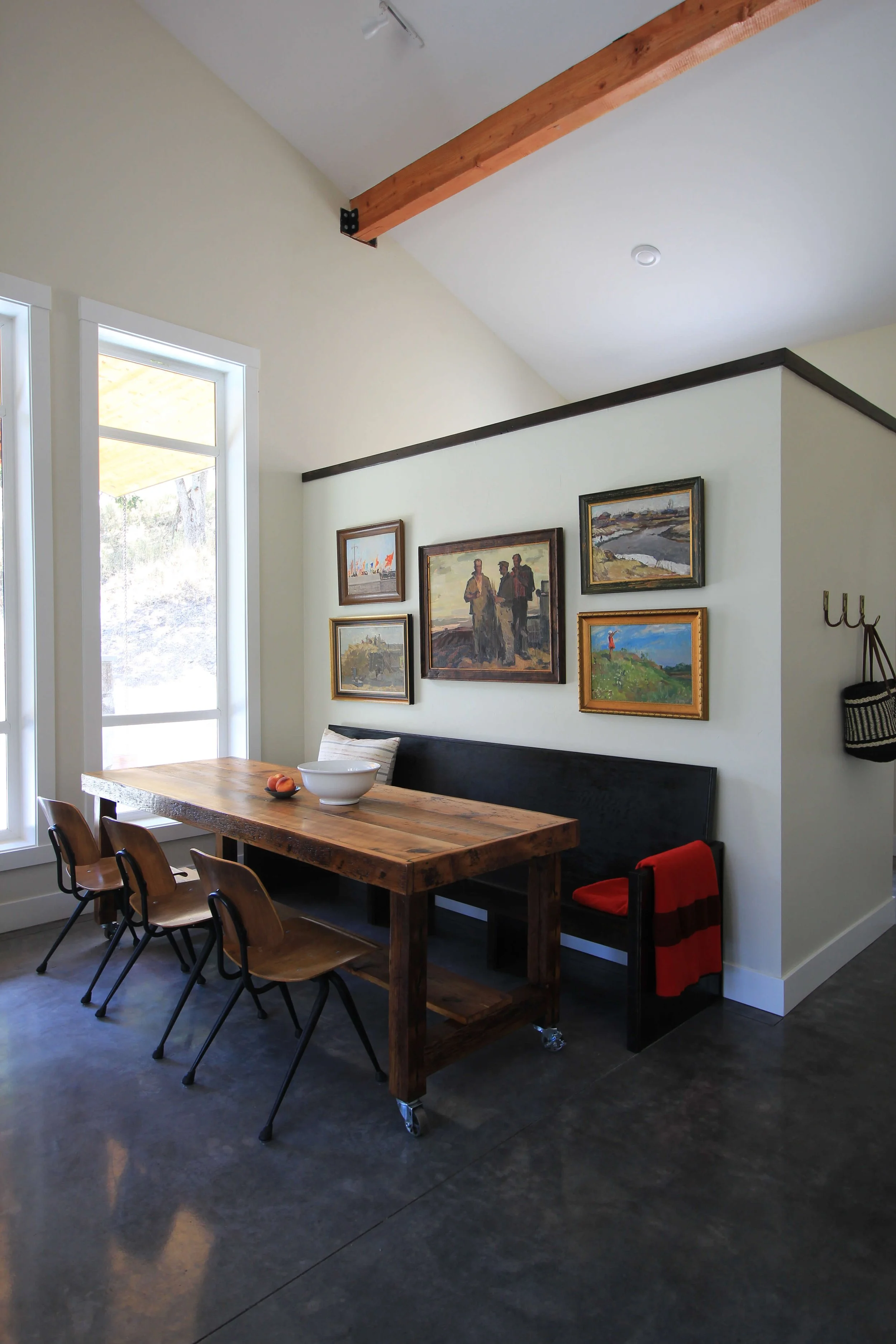A dining area with a wooden table, six chairs, a black bench, and wall art. There are two large windows on the left, and the ceiling has a wooden beam. The floor is dark-colored, and the walls are painted light.
