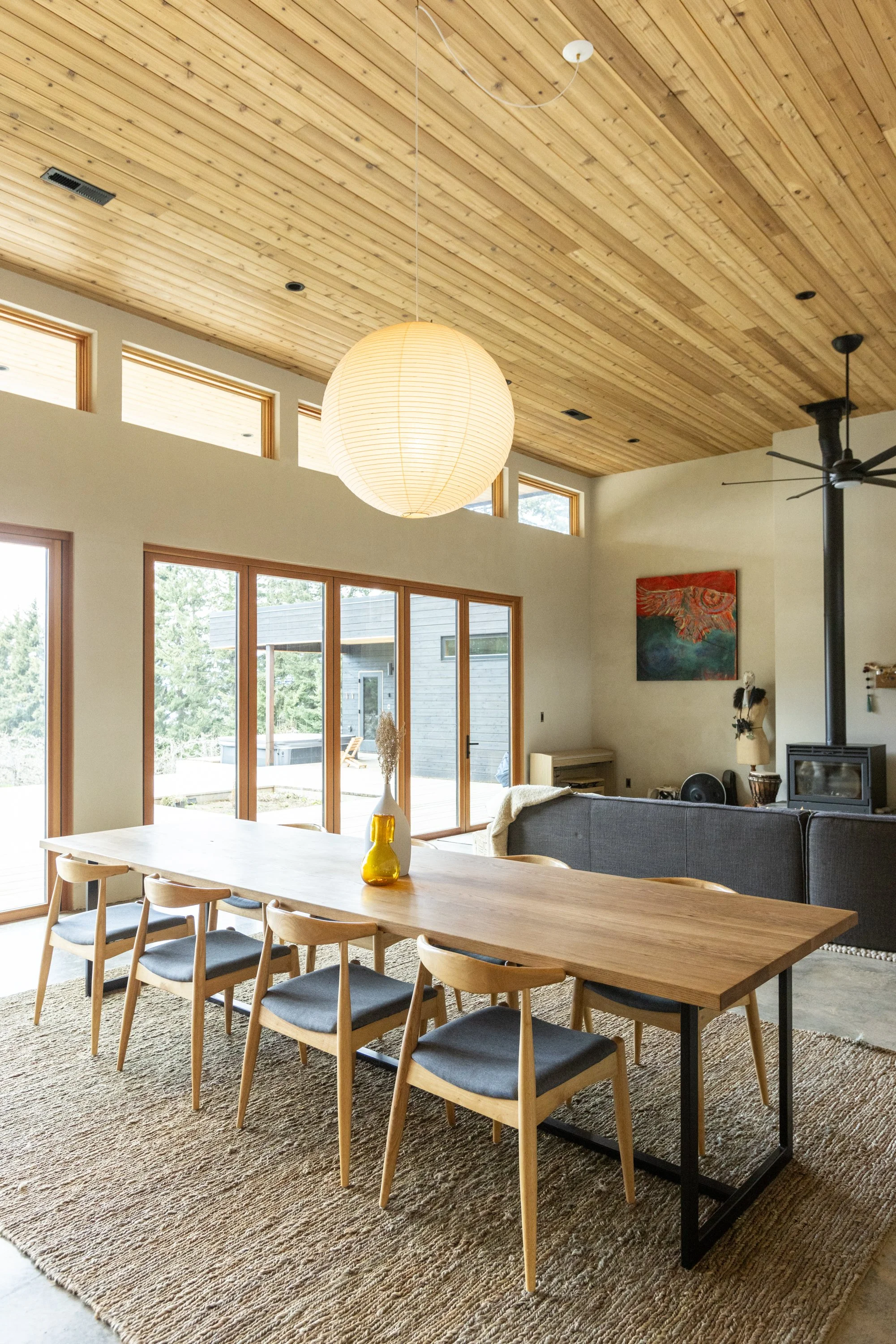 Modern dining and living area with wooden ceiling, large windows, and a hanging paper lantern light fixture. Dining table with chairs on a textured rug, and a living room with a gray sofa, art on the wall, and a stacked wood stove.